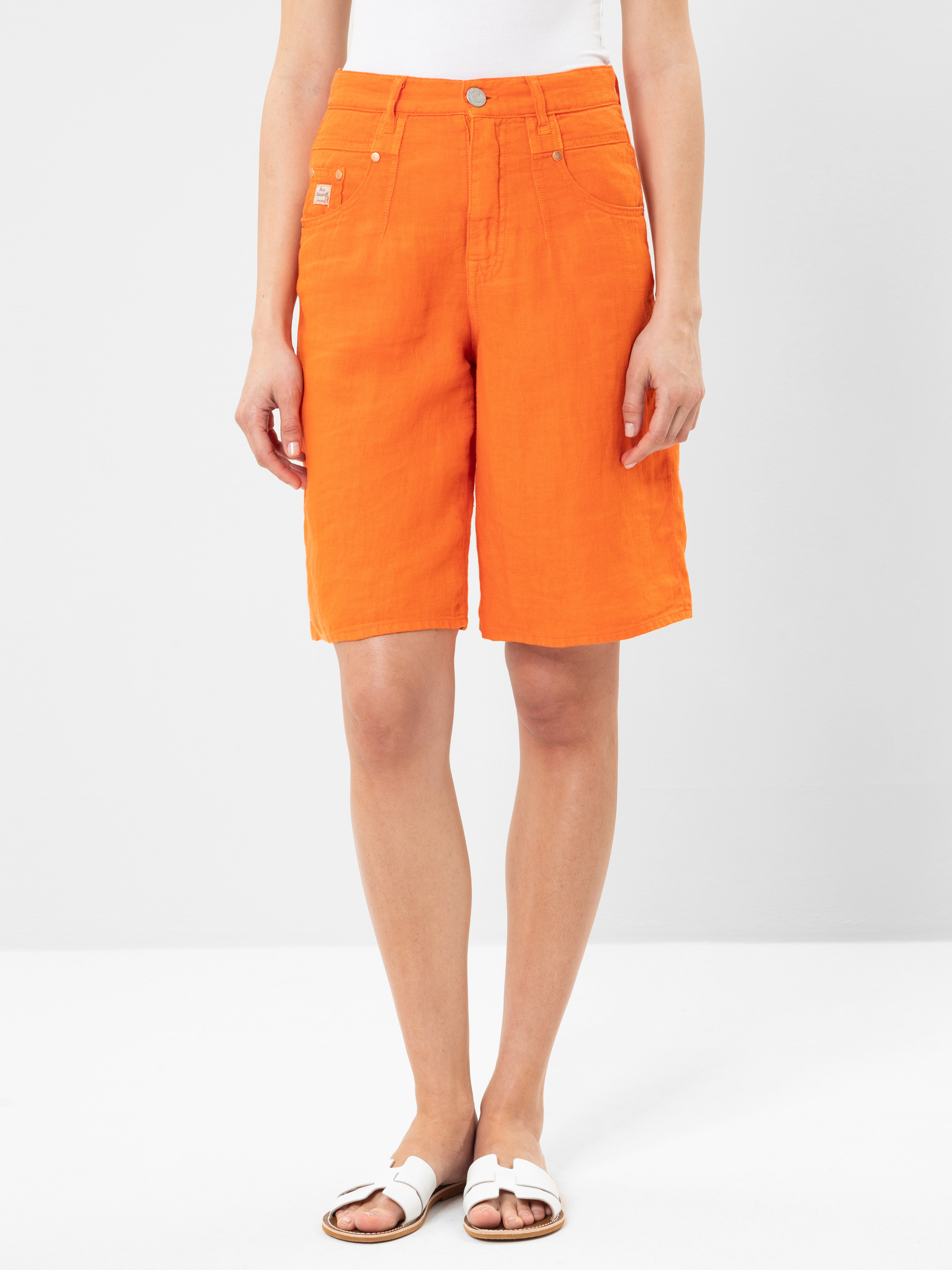 Eine Person, die leuchtend orangefarbene knielange Shorts, ein weißes ärmelloses Oberteil und weiße Sandalen trägt, steht vor einem schlichten hellgrauen Hintergrund.