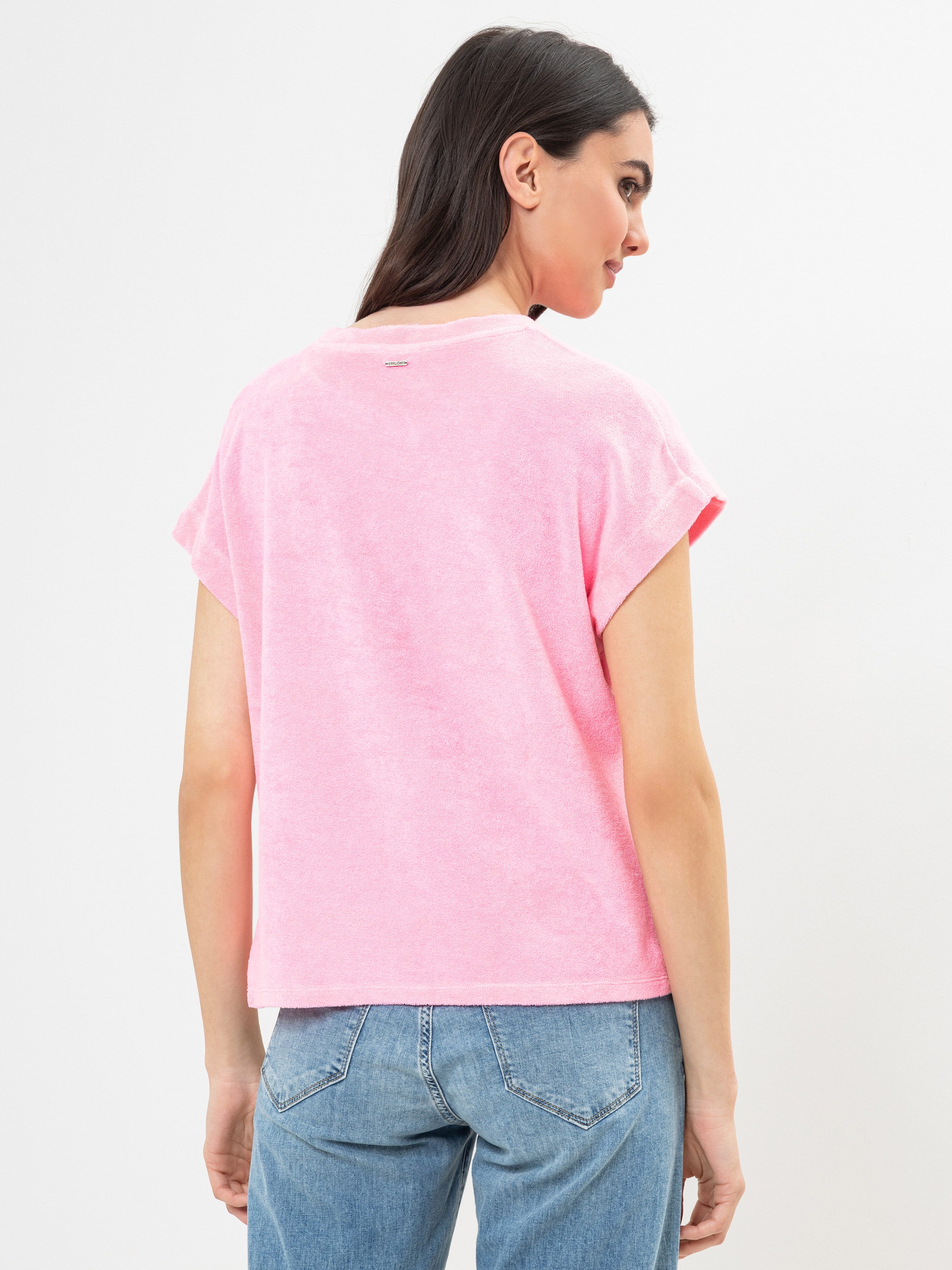 Eine Frau mit langen dunklen Haaren trägt ein locker sitzendes rosa kurzärmeliges Oberteil und blaue Jeans. Sie steht mit dem Gesicht zur Seite und zeigt die Rückseite ihres Outfits vor einem schlichten weißen Hintergrund.