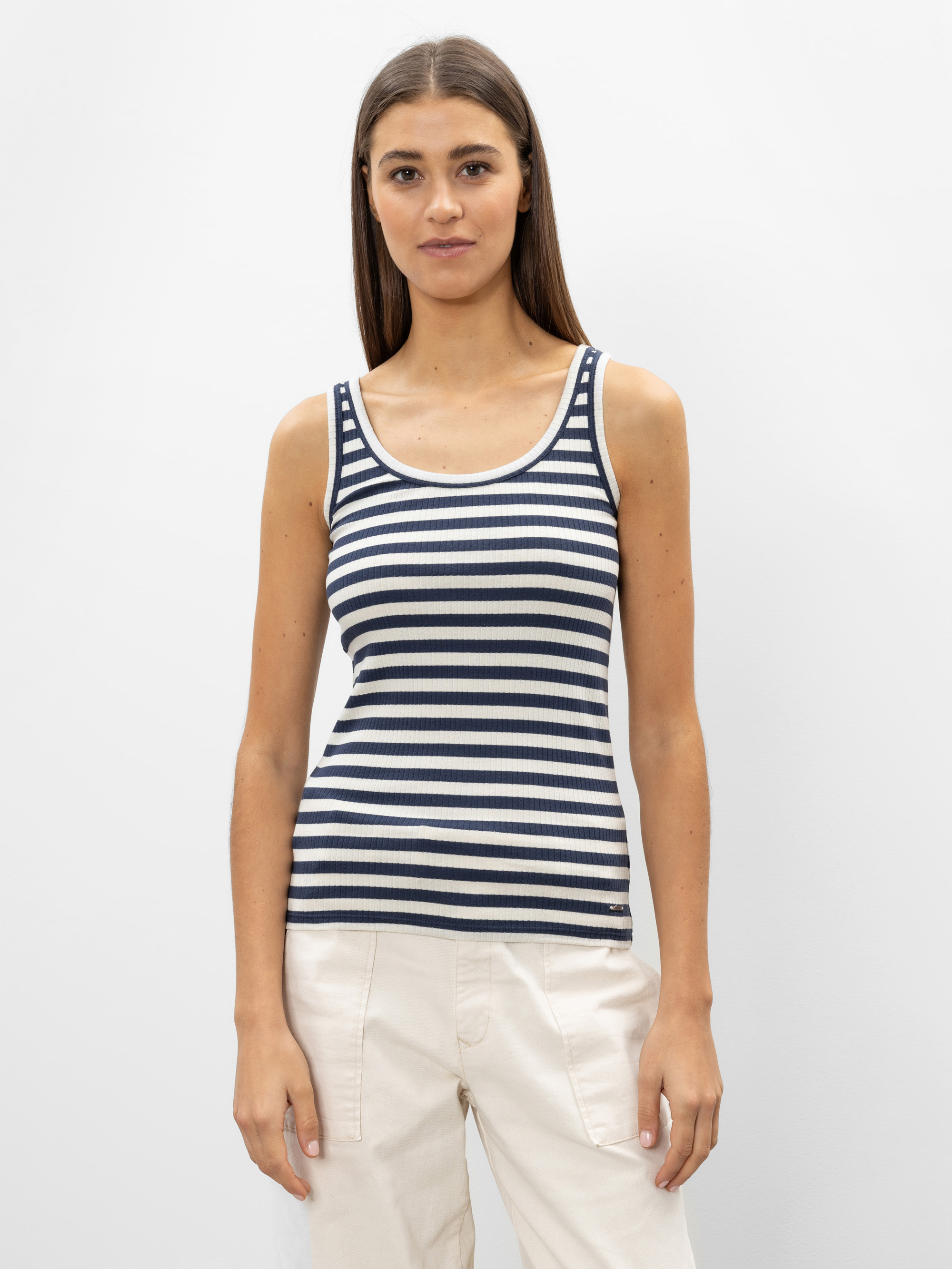 Eine Frau mit glattem braunem Haar trägt ein marineblau-weiß gestreiftes ärmelloses Tank-Top und eine hellbeige Hose vor einem schlichten weißen Hintergrund.