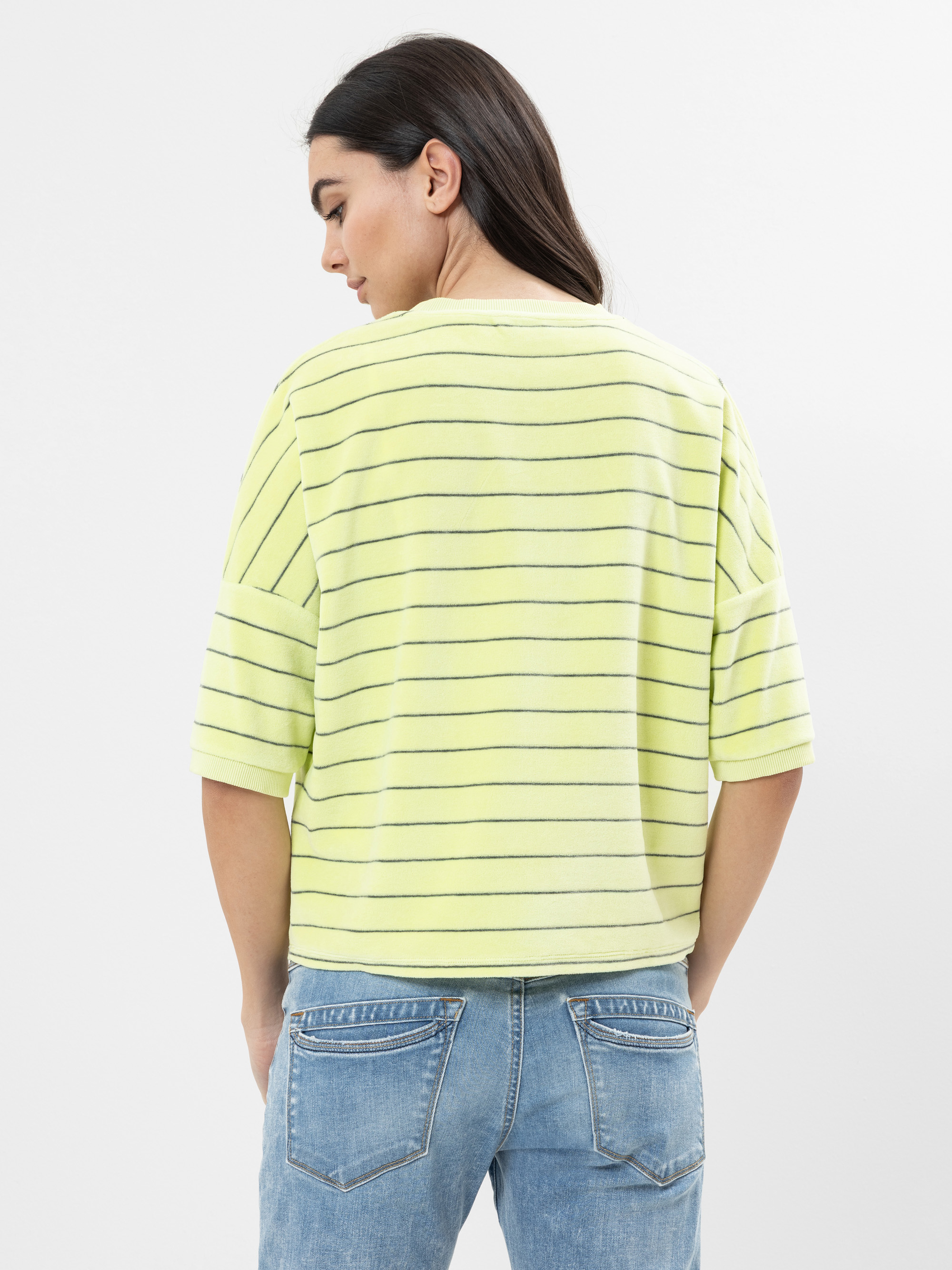 Eine Frau mit langen dunklen Haaren steht mit dem Rücken zur Kamera und trägt ein hellgelbes kurzärmeliges Oberteil mit dünnen Querstreifen und eine blaue Jeans. Der Hintergrund ist einfarbig und weiß.