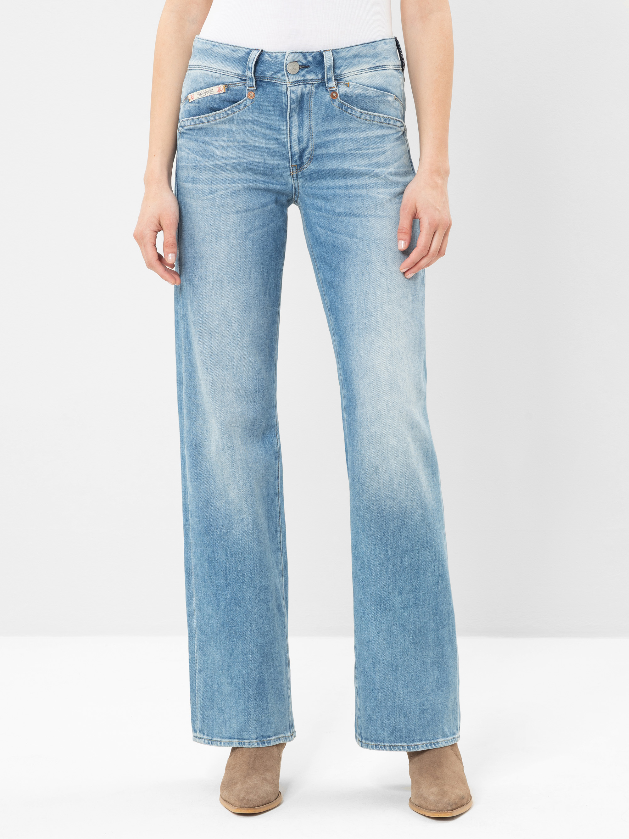 Eine Person, die hellblaue, hochtaillierte Jeans mit weitem Bein, ein weißes Oberteil und hellbraune Stiefeletten trägt, steht vor einem schlichten weißen Hintergrund. Das Bild zeigt den Körper von den Schultern bis zu den Füßen.