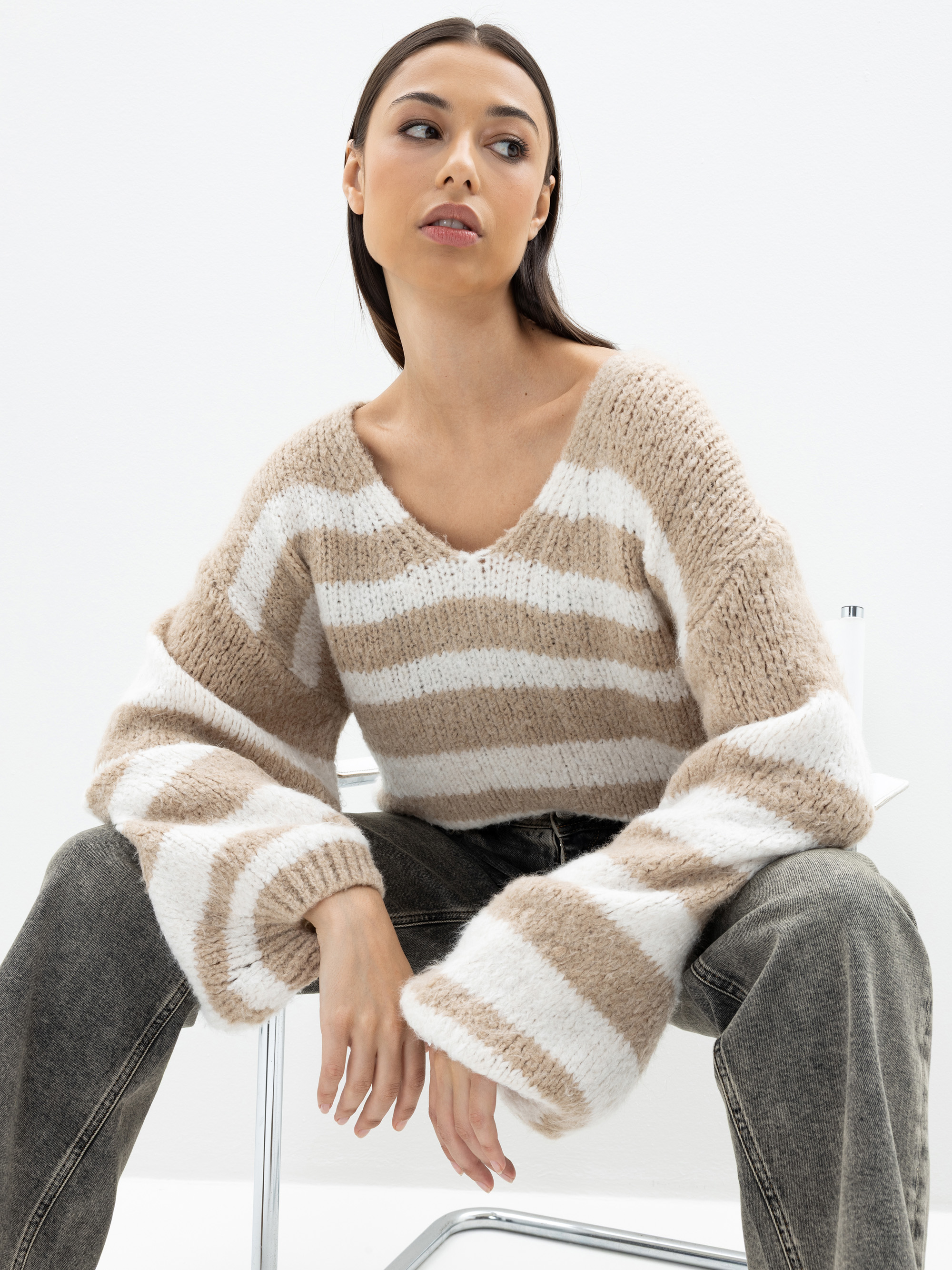 Eine Frau mit langen dunklen Haaren sitzt auf einem weißen Stuhl, trägt einen beige-weiß gestreiften Strickpullover mit weiten Ärmeln und graue Jeans und blickt leicht zur Seite vor einem schlichten weißen Hintergrund.