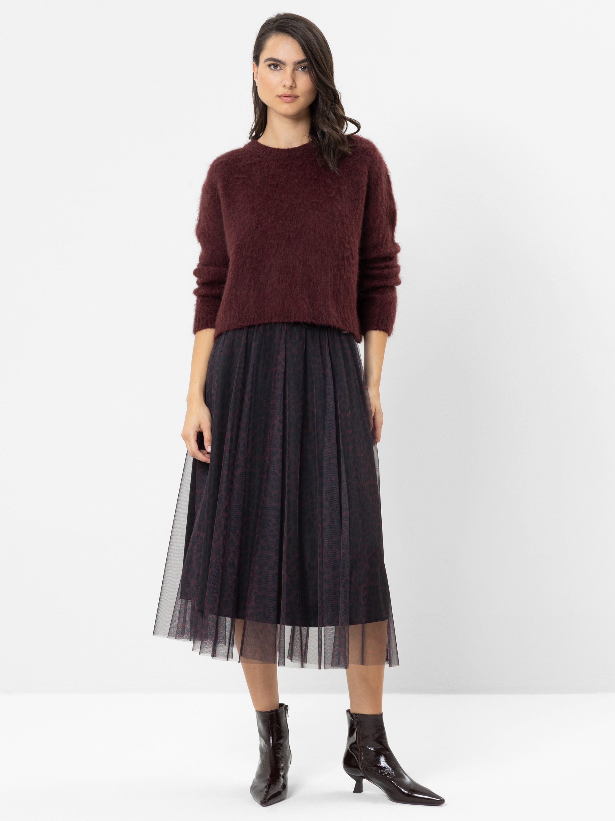 Eine Frau steht vor einem weißen Hintergrund und trägt einen burgunderroten, flauschigen Pullover, einen schwarzen Midi-Tüllrock mit dezentem Muster sowie glänzende Stiefeletten. Sie hat dunkles, welliges Haar und einen neutralen Gesichtsausdruck.