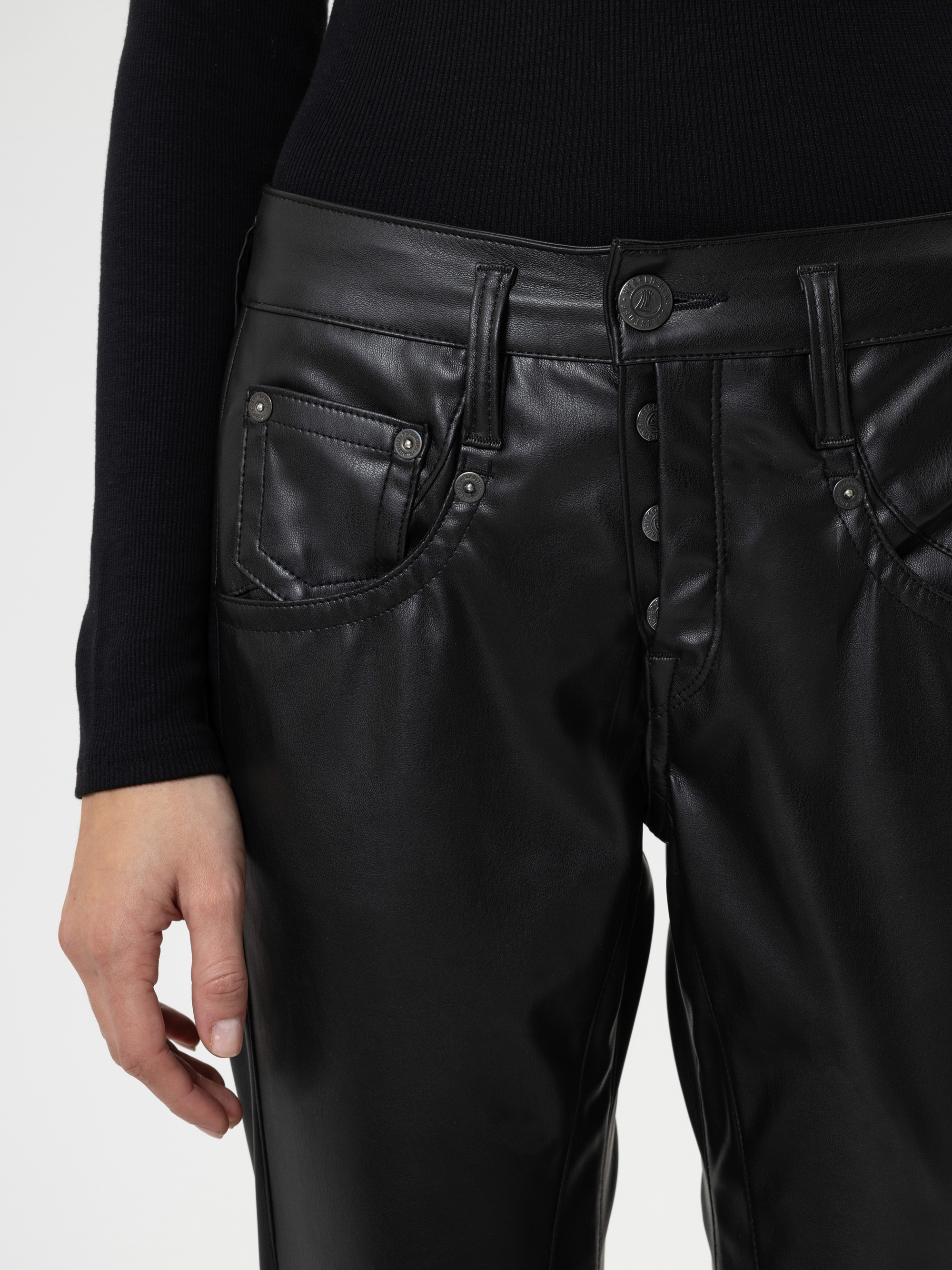 Eine Person, die eine schwarze Kunstlederhose und ein schwarzes langärmeliges Oberteil trägt und ihre linke Hand an der Seite ruhen lässt. Der Schwerpunkt des Bildes liegt auf der Taille und den Hüften.