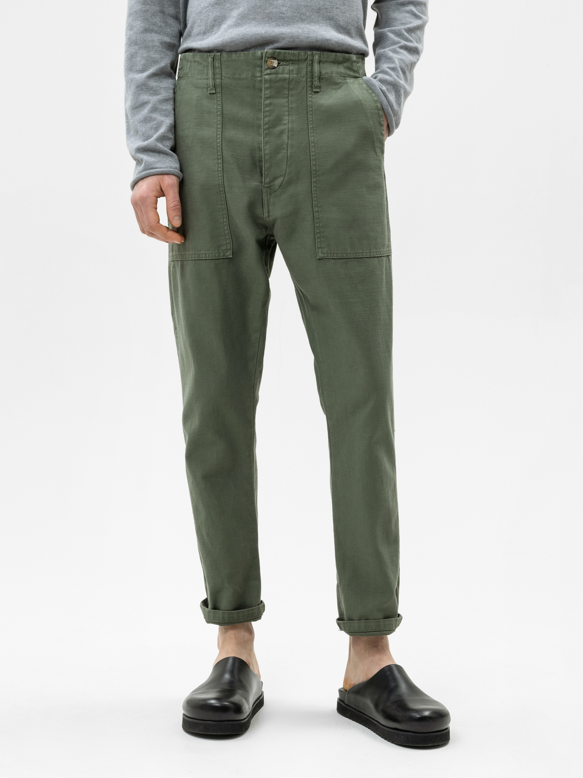 Eine Person, die eine olivgrüne, hoch taillierte Hose mit großen Vordertaschen, ein graues, langärmeliges Hemd und schwarze Slipper trägt, steht vor einem schlichten weißen Hintergrund.