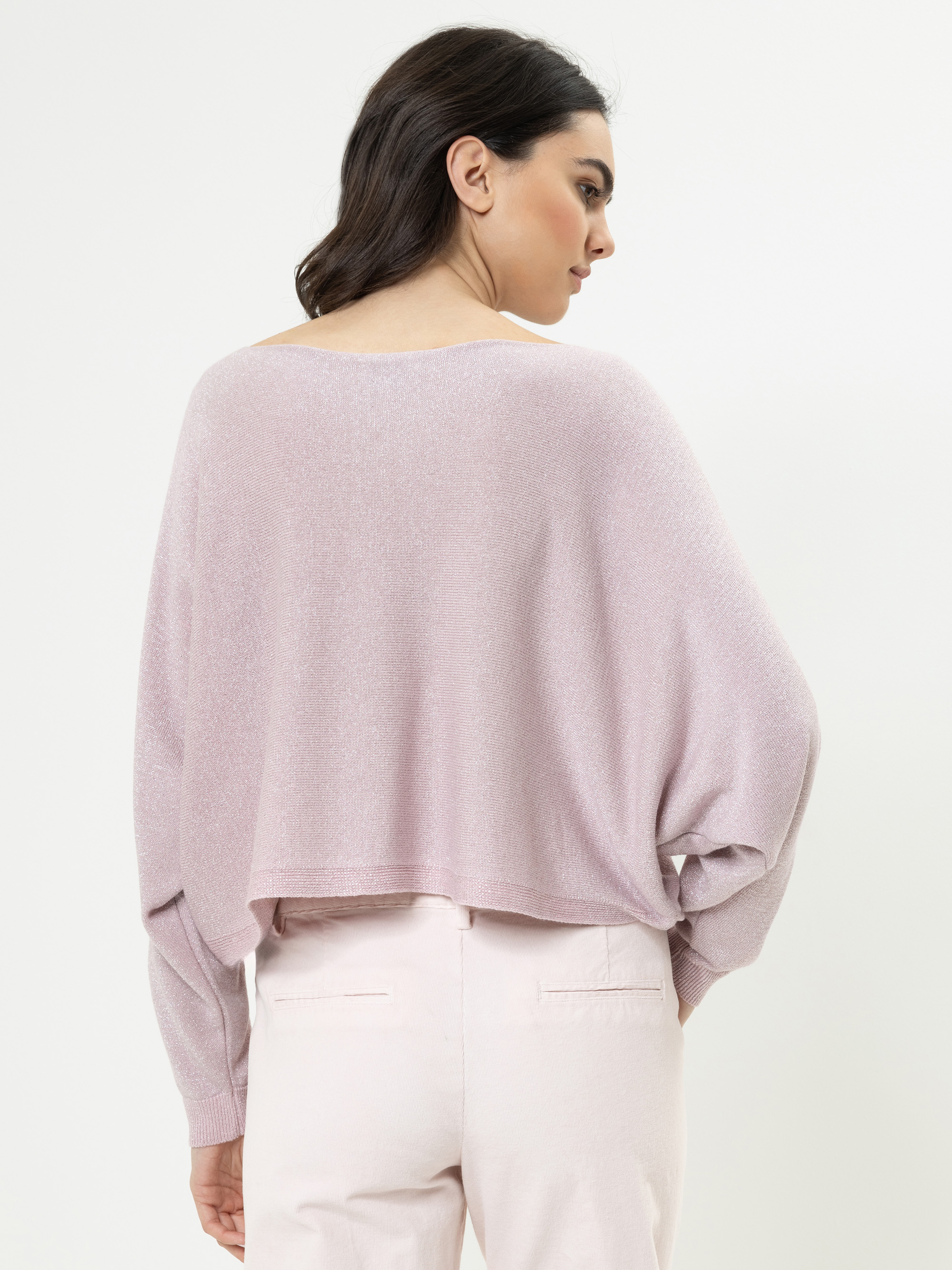 Eine Frau mit dunklem Haar, das in lockeren Wellen frisiert ist, ist von hinten zu sehen. Sie trägt einen hellvioletten, locker sitzenden Pullover und eine blassrosa Hose vor einem schlichten weißen Hintergrund.