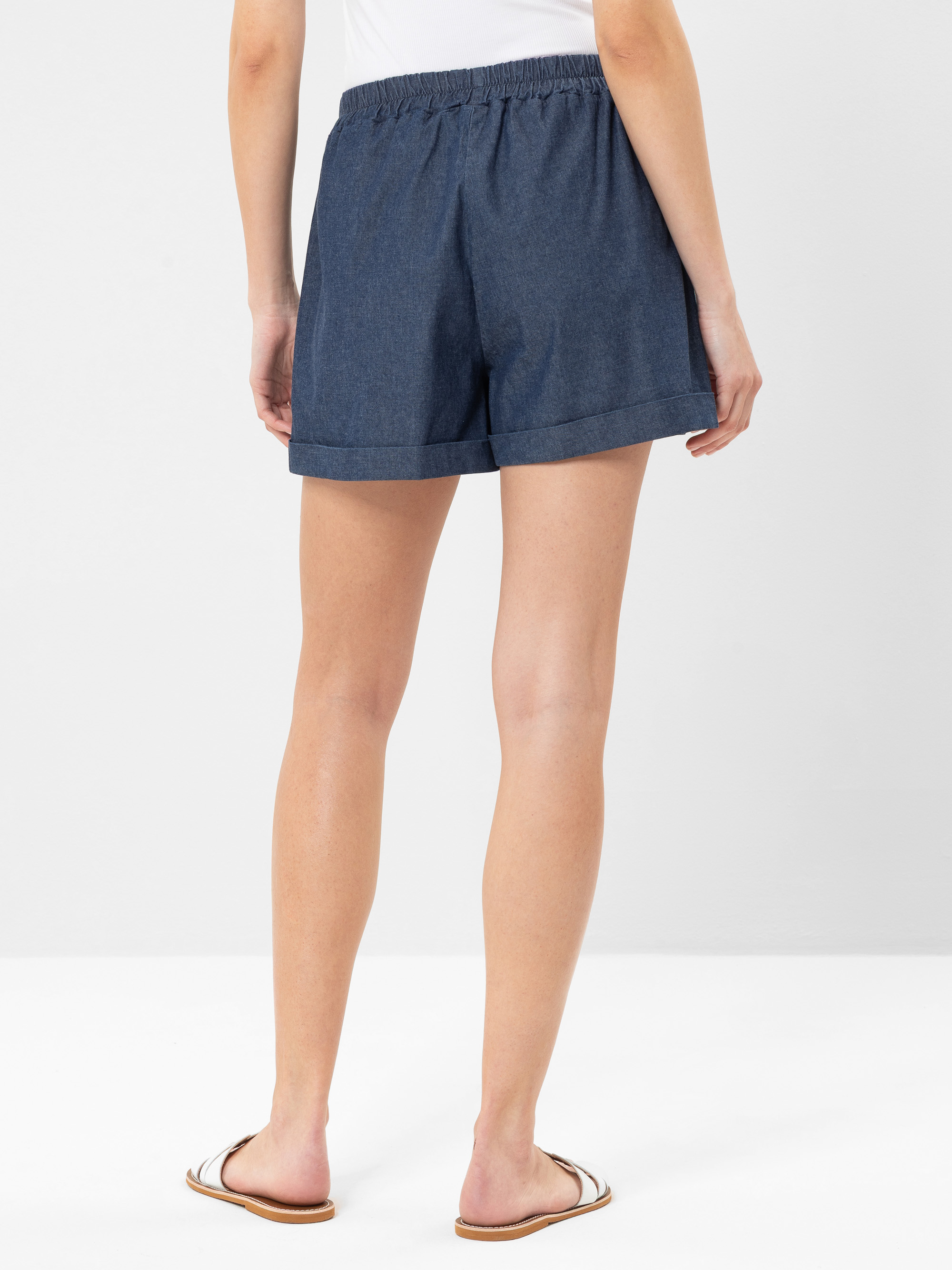 Eine Person, die ein weißes Oberteil, dunkelblaue Jeansshorts und weiße Flip-Flops trägt, steht mit dem Gesicht nach hinten und zeigt die Rückseite der Shorts vor einem schlichten weißen Hintergrund.