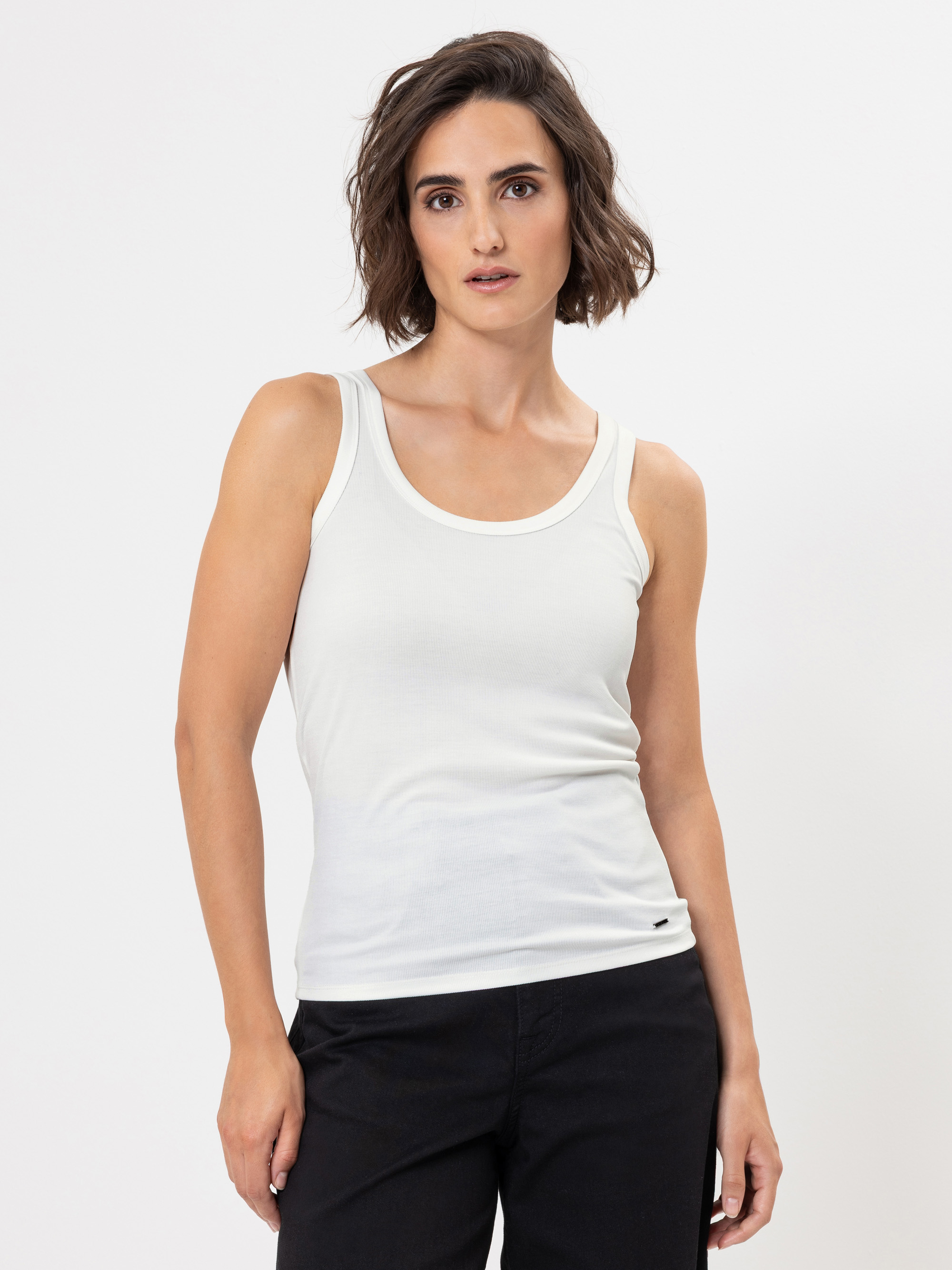 Eine Frau mit kurzen braunen Haaren, die ein weißes ärmelloses Tank-Top und eine schwarze Hose trägt, steht vor einem schlichten weißen Hintergrund und schaut mit neutralem Blick direkt in die Kamera.