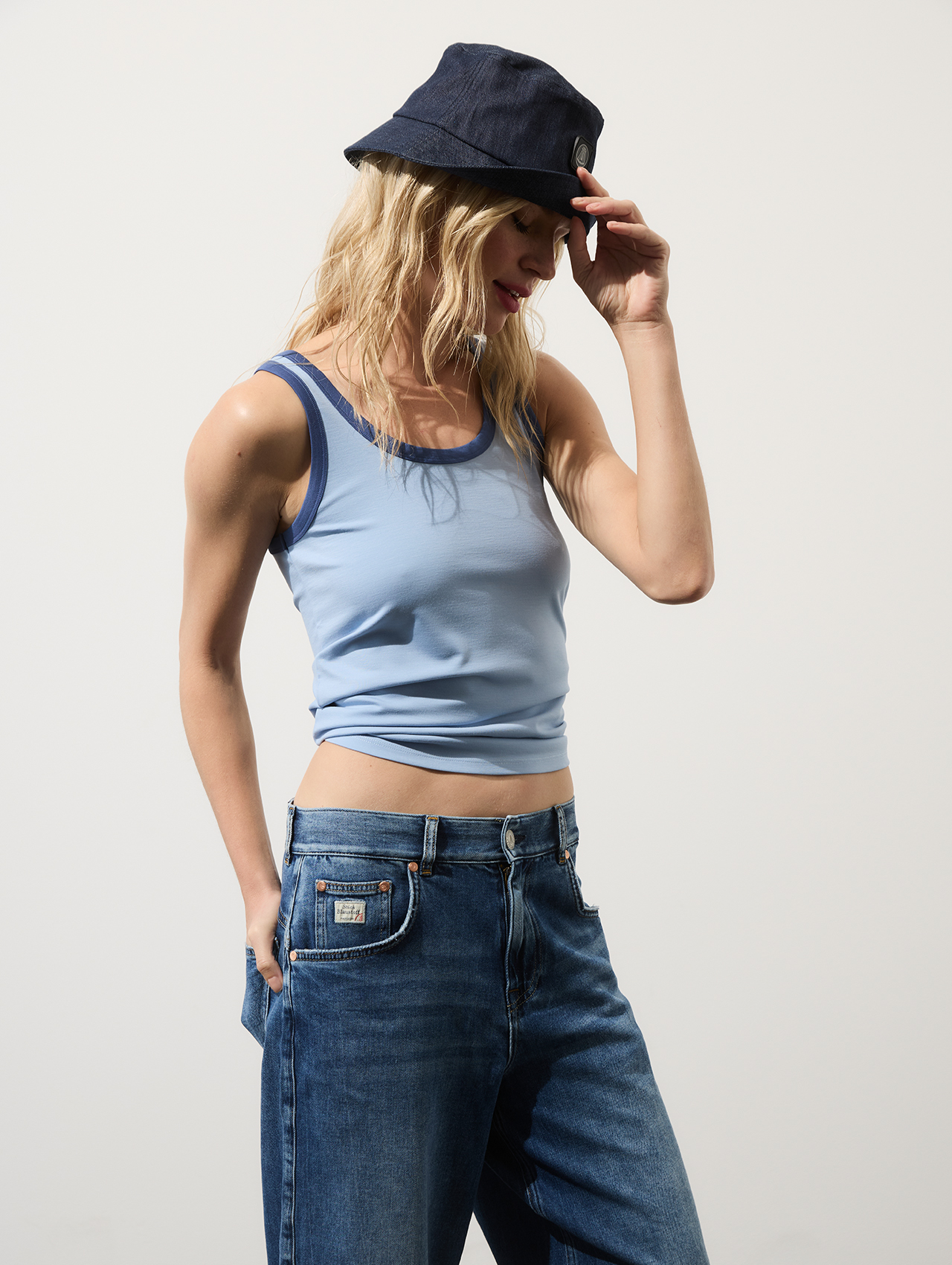 Eine Frau mit einem marineblauen Bucket-Hat, einem hellblauen Tanktop und einer blauen Jeans steht vor einem schlichten hellen Hintergrund, blickt nach unten und berührt mit einer Hand die Krempe ihres Hutes.