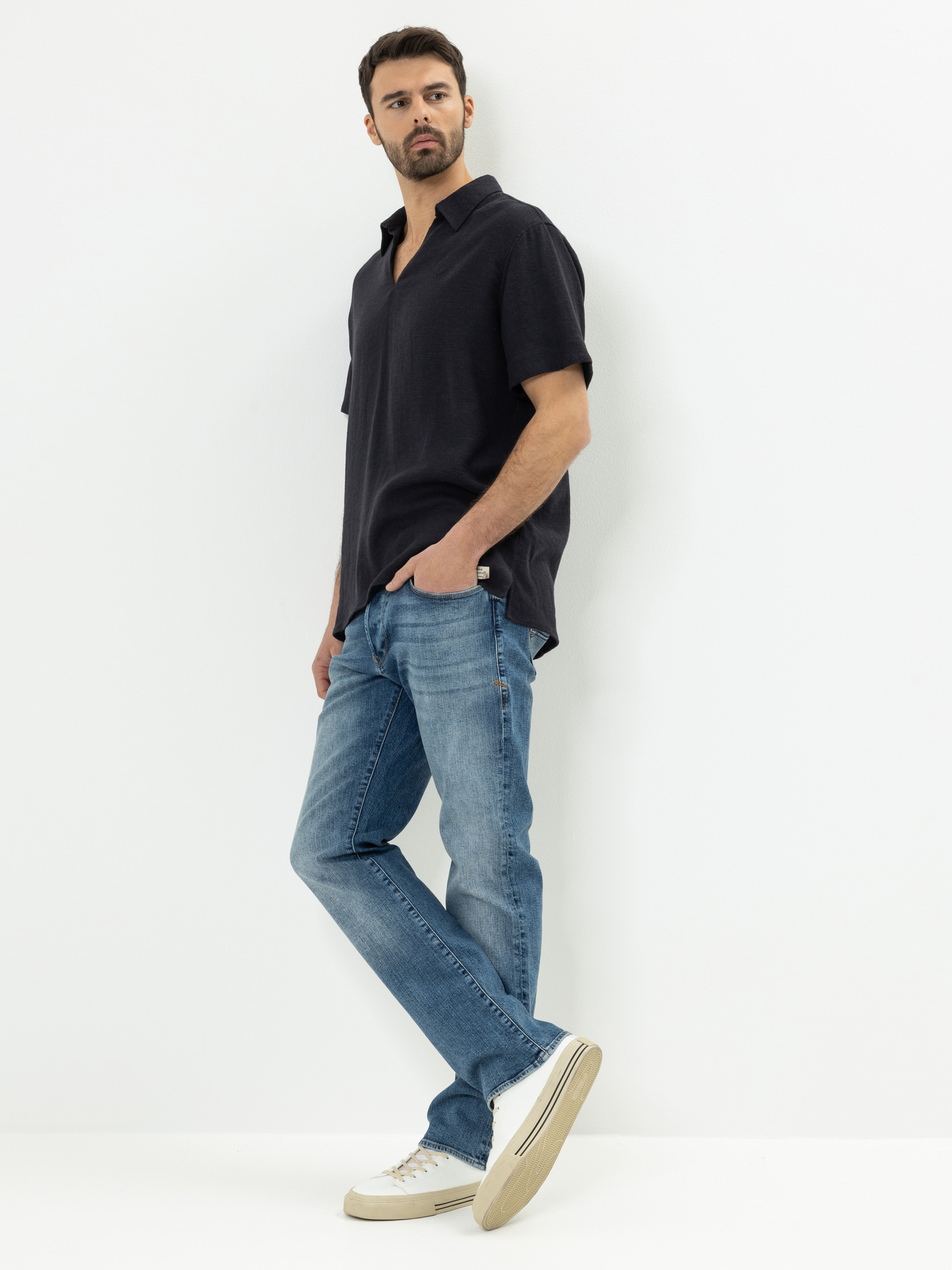 Ein Mann steht vor einem weißen Hintergrund und trägt ein dunkles, kurzärmeliges Hemd, hellblaue Jeans und weiße Turnschuhe. Eine Hand steckt in seiner Tasche, während er zur Seite schaut.