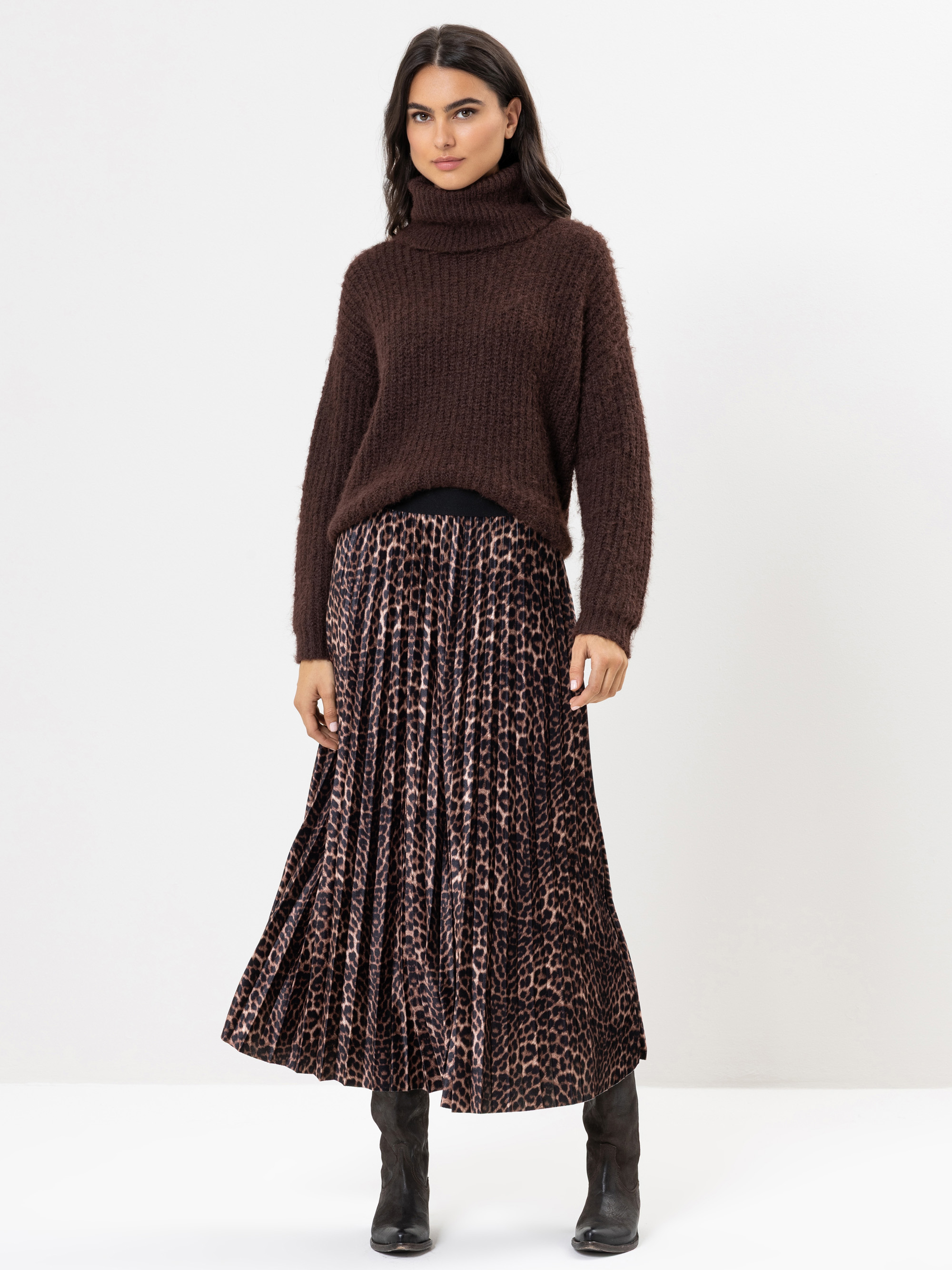 Eine Frau steht vor einem weißen Hintergrund und trägt einen braunen Rollkragenpullover, einen gefalteten Maxirock mit Leopardenmuster und schwarze Stiefel. Sie hat langes dunkles Haar und einen neutralen Gesichtsausdruck.