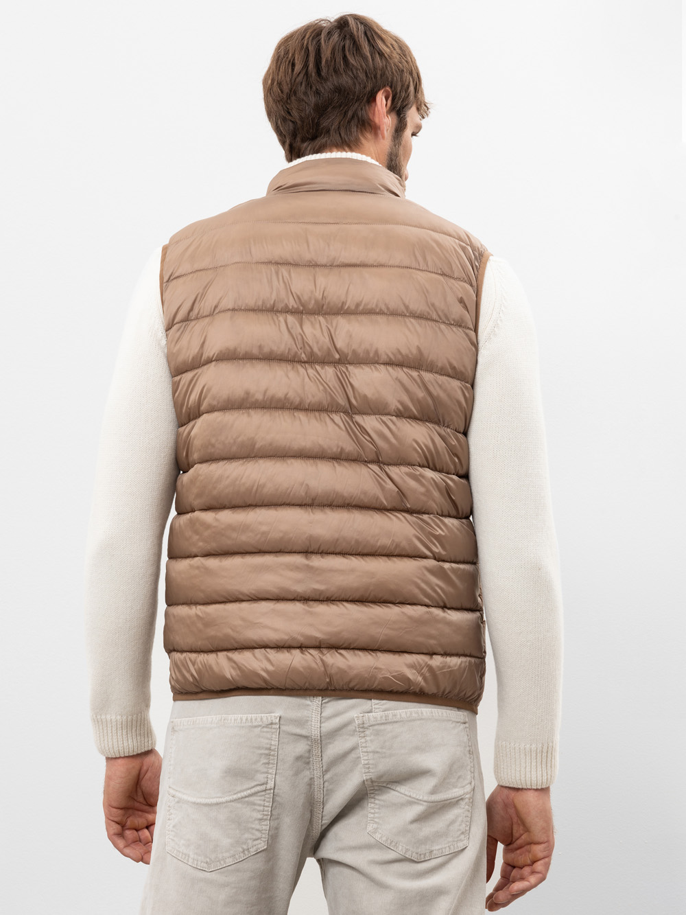 Ein Mann steht mit dem Rücken zur Kamera und trägt eine hellbraune Steppweste über einem cremefarbenen Pullover und hellgraue Jeans vor einem schlichten hellen Hintergrund.