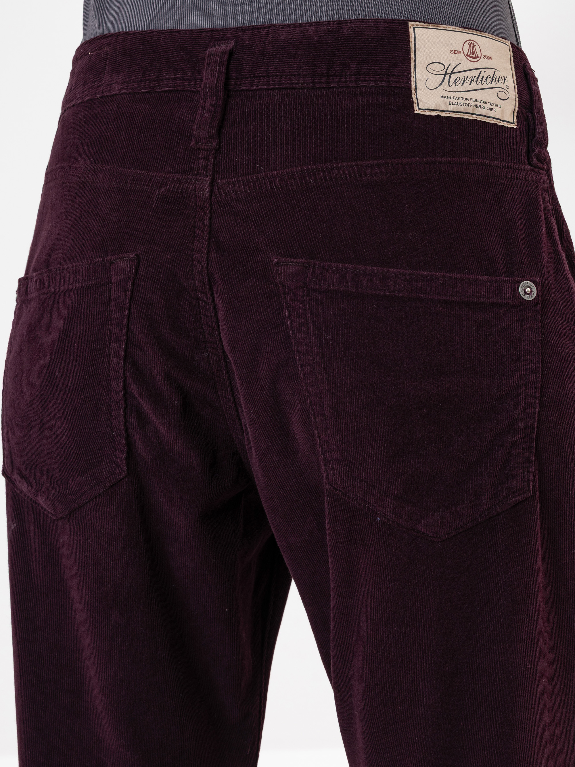 Nahaufnahme des Rückens einer Person, die eine dunkelviolette Cordhose mit zwei Gesäßtaschen und einem beigen Herrlicher-Markenetikett am Bund trägt.