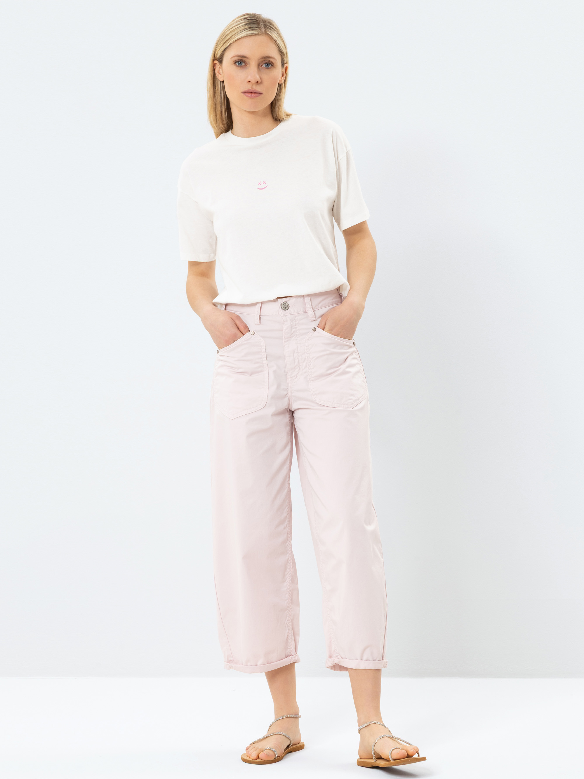 Eine Frau steht vor einem schlichten weißen Hintergrund und trägt ein weißes T-Shirt, eine hellrosa Hose mit weitem Bein und beigefarbene Sandalen. Sie hat glattes blondes Haar und ihre Hände stecken in den Taschen.