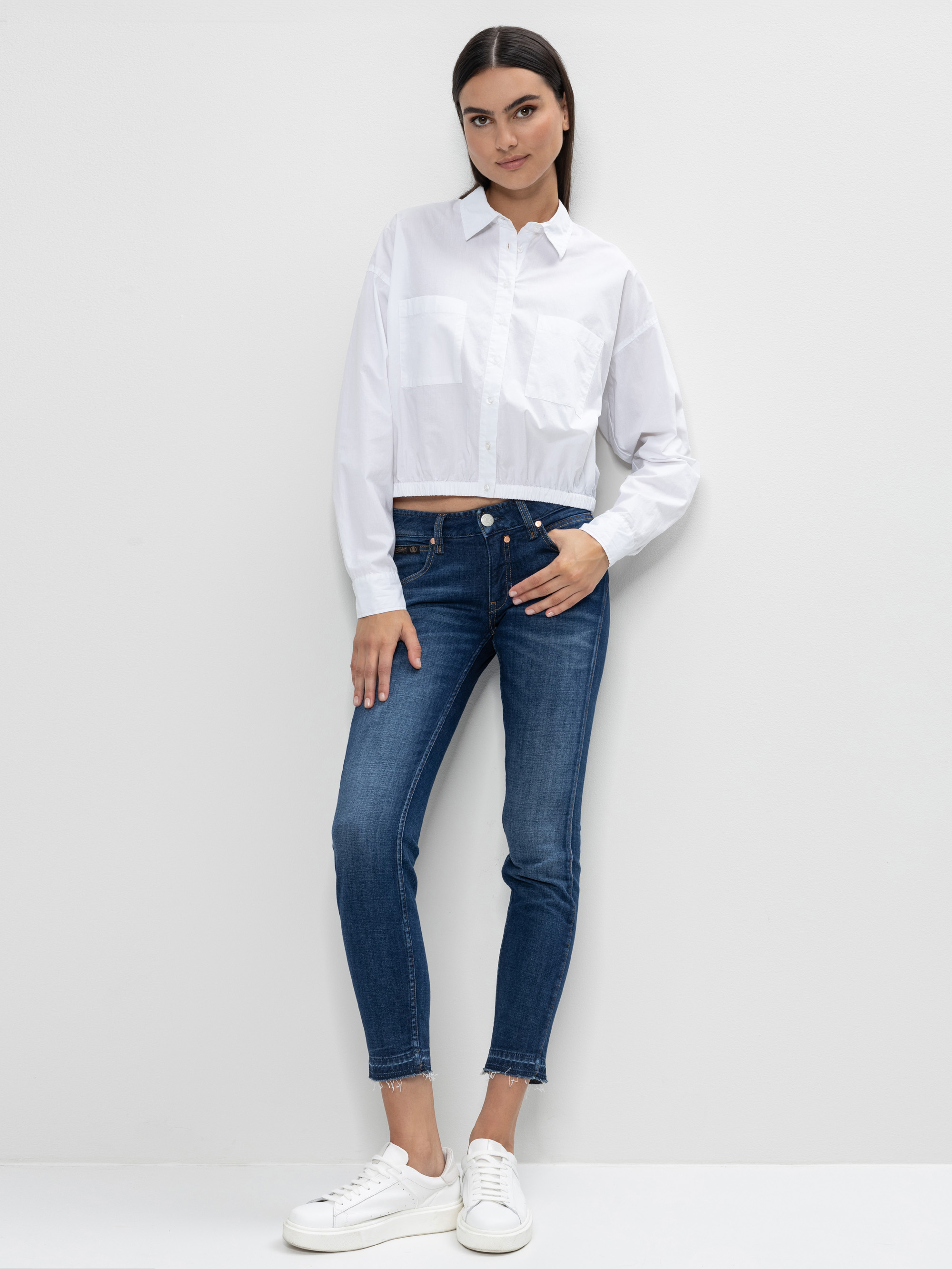 Eine Frau steht vor einer schlichten weißen Wand und trägt ein weißes Button-up-Hemd, blaue Skinny-Jeans und weiße Turnschuhe. Sie hat glattes dunkles Haar und eine entspannte, selbstbewusste Pose mit einer Hand in der Tasche.