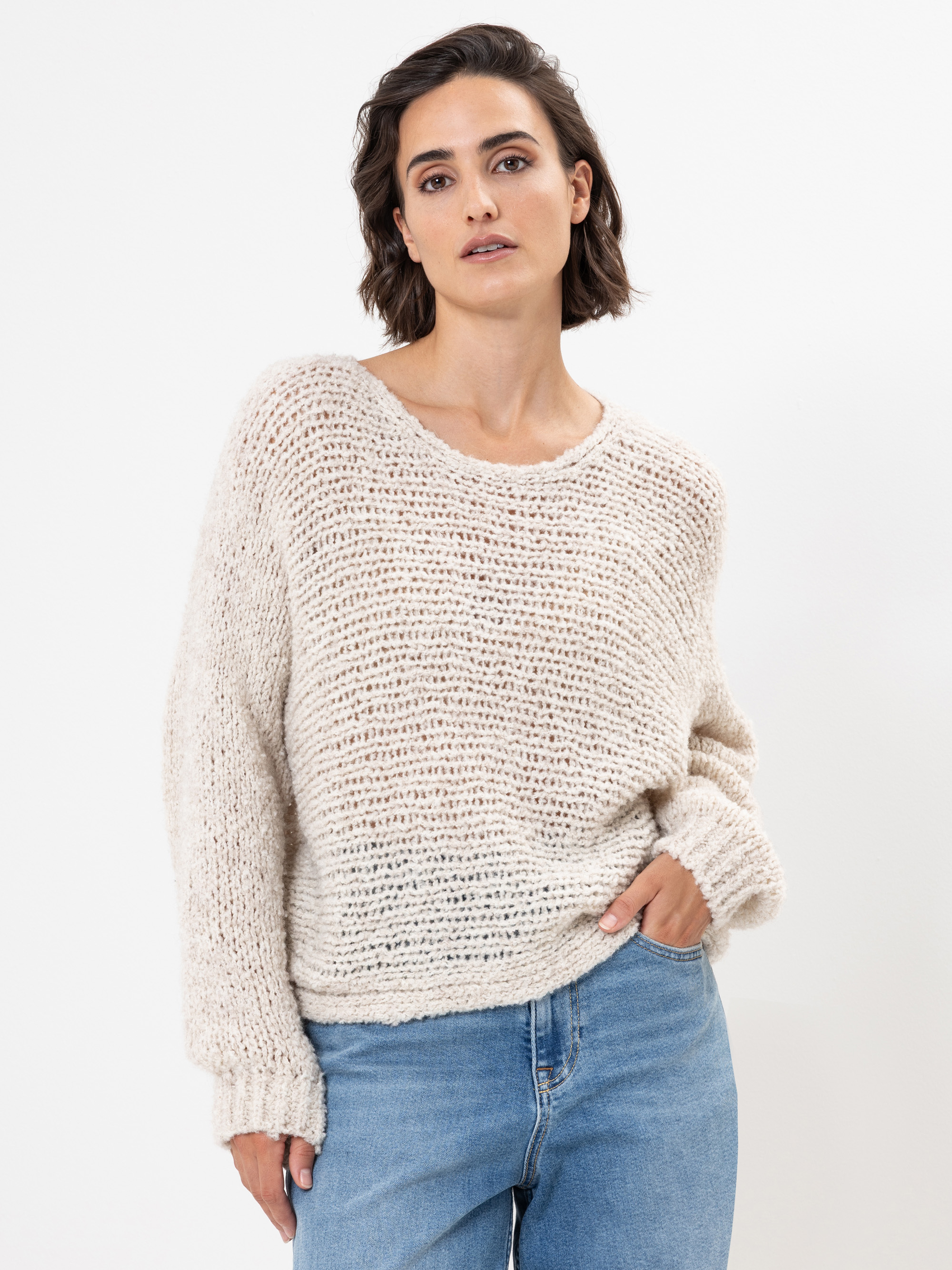 Eine Frau mit kurzen braunen Haaren trägt einen lockeren, cremefarbenen Strickpullover und hellblaue Jeans und steht mit entspanntem Gesichtsausdruck vor einem schlichten weißen Hintergrund.