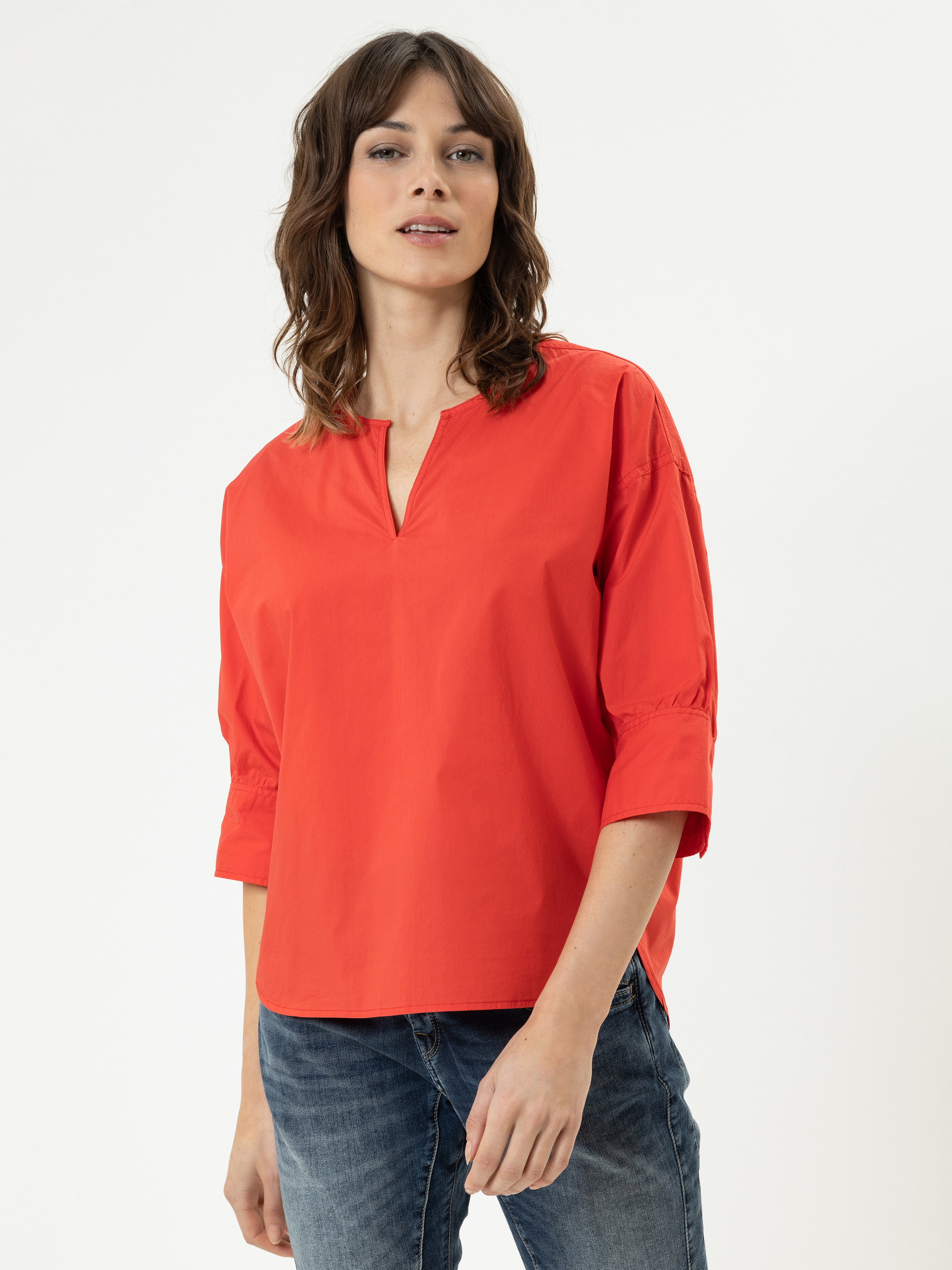 Eine Frau mit gewelltem braunem Haar trägt ein leuchtend rotes, locker sitzendes Oberteil mit dreiviertel Ärmeln und gekerbtem Ausschnitt, gepaart mit blauen Jeans, vor einem schlichten weißen Hintergrund.