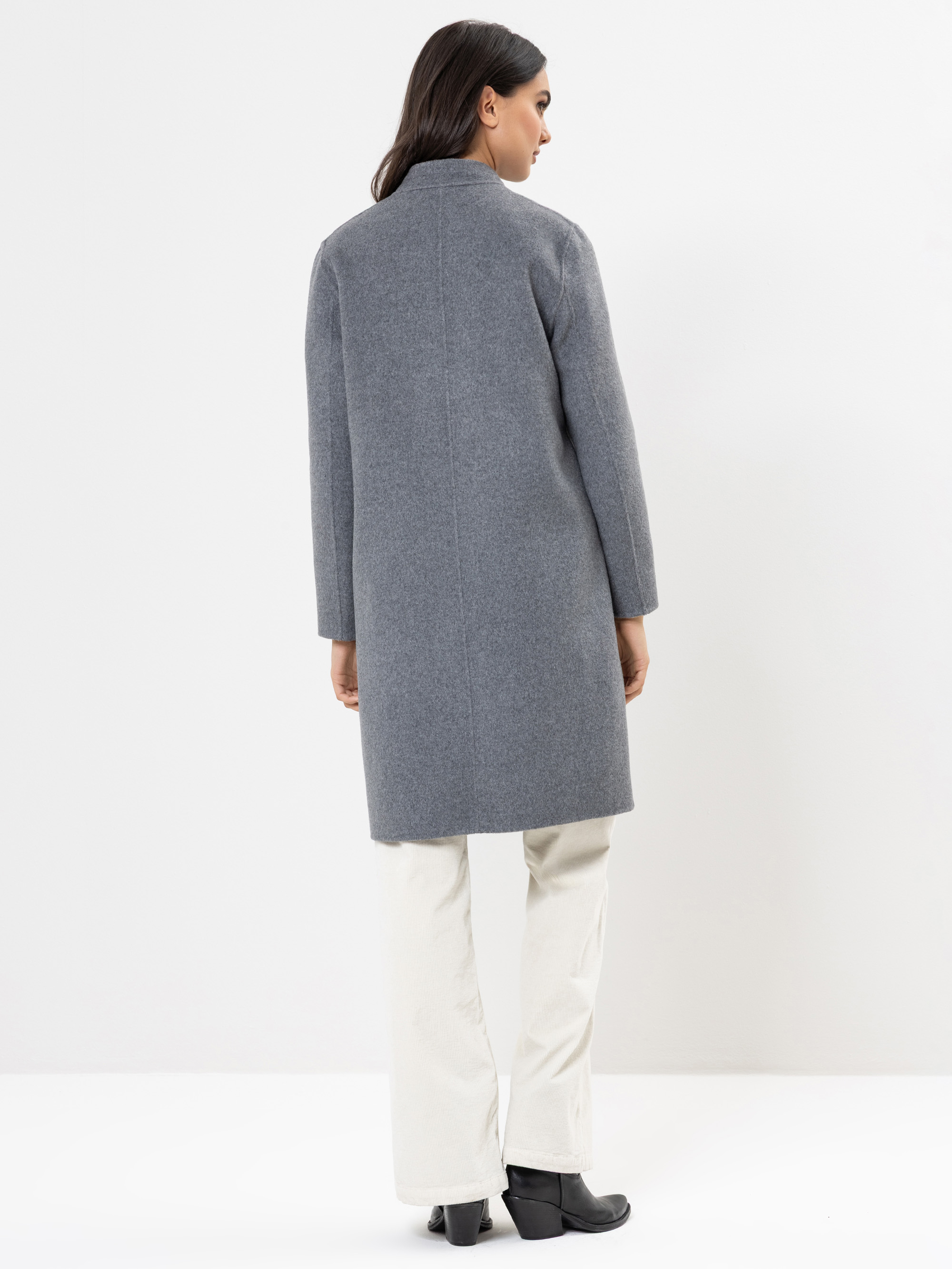 Eine Frau steht in einem langen grauen Mantel, einer weißen Hose und schwarzen Stiefeln vor einem schlichten weißen Hintergrund.