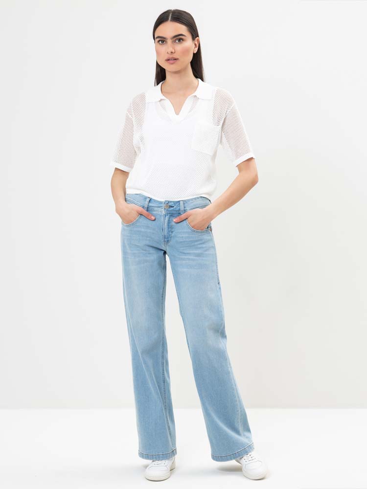 Eine Frau steht vor einem weißen Hintergrund und trägt ein weißes kurzärmeliges Hemd mit Netzärmeln, hellblaue Jeans mit weitem Bein und weiße Turnschuhe. Sie hat die Hände in den Taschen und trägt langes, glattes dunkles Haar.