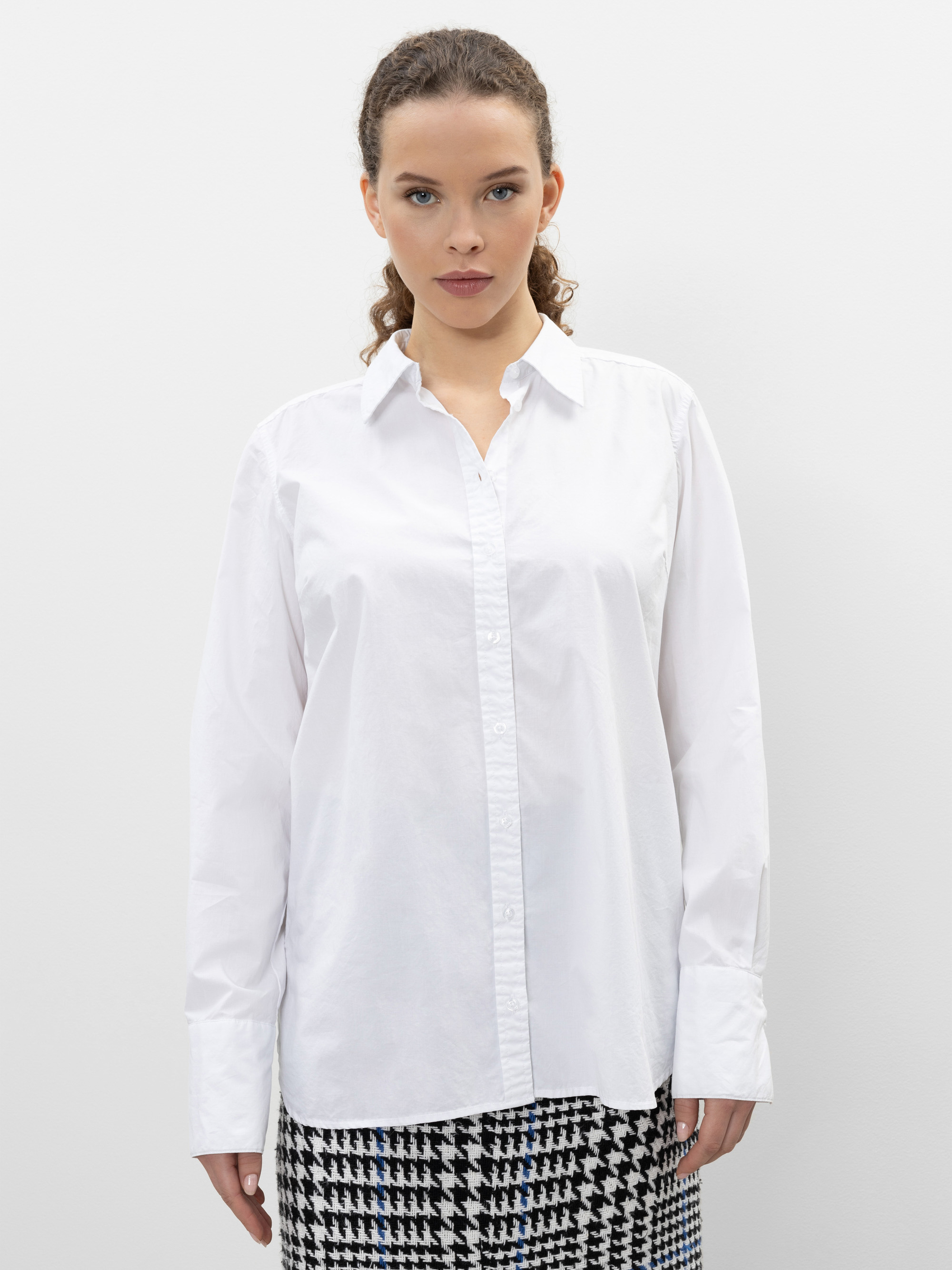 Eine Frau mit lockigem Haar trägt ein weißes Button-up-Hemd und einen schwarz-weißen Hahnentritt-Rock und steht vor einem schlichten weißen Hintergrund.