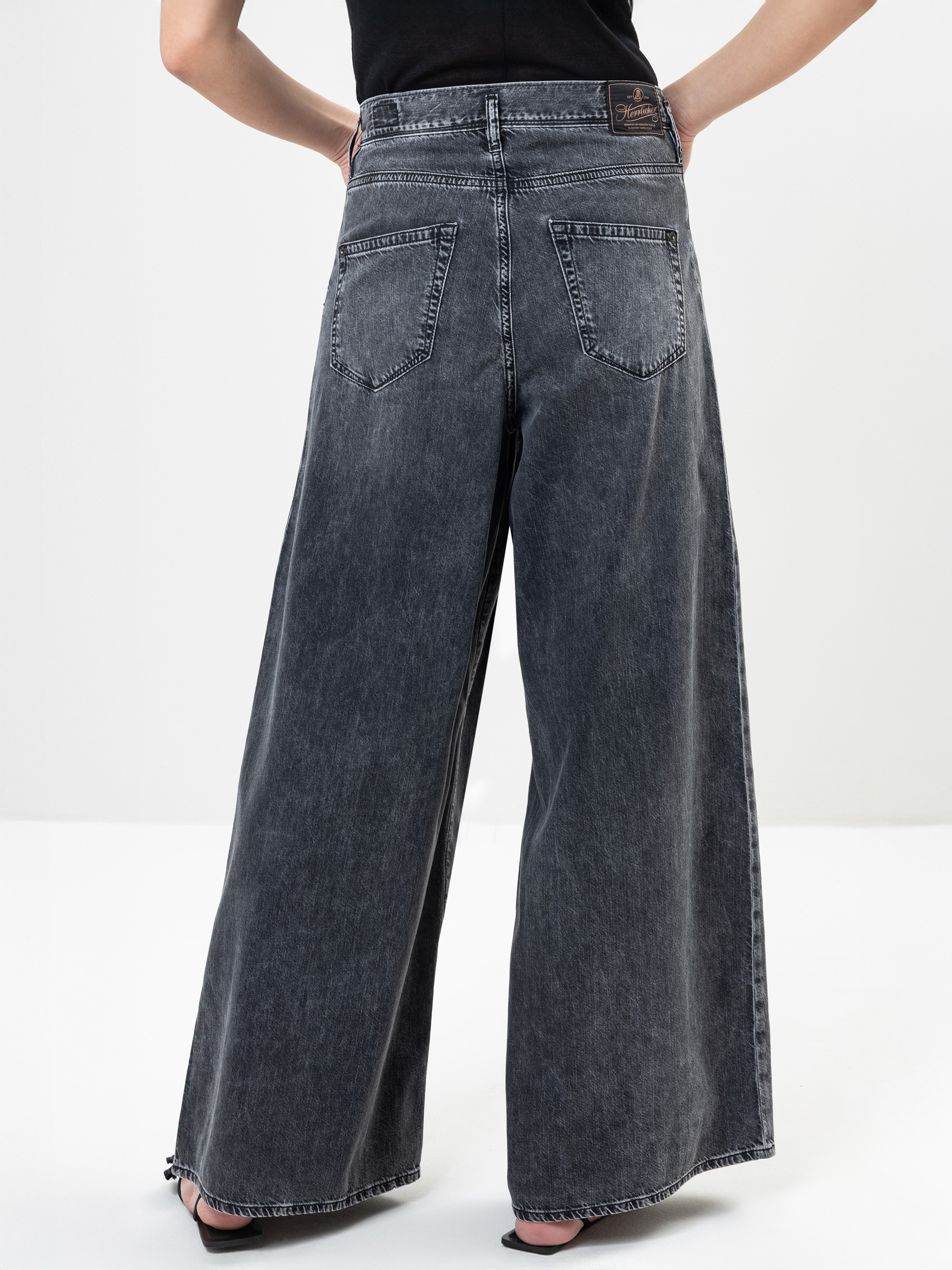 Eine Person, die eine schwarze, verblichene Jeans mit weitem Bein und hoher Taille und ausgefransten Säumen trägt, von hinten gesehen. Die Jeans hat zwei Gesäßtaschen und die Person steht mit den Händen auf den Hüften.