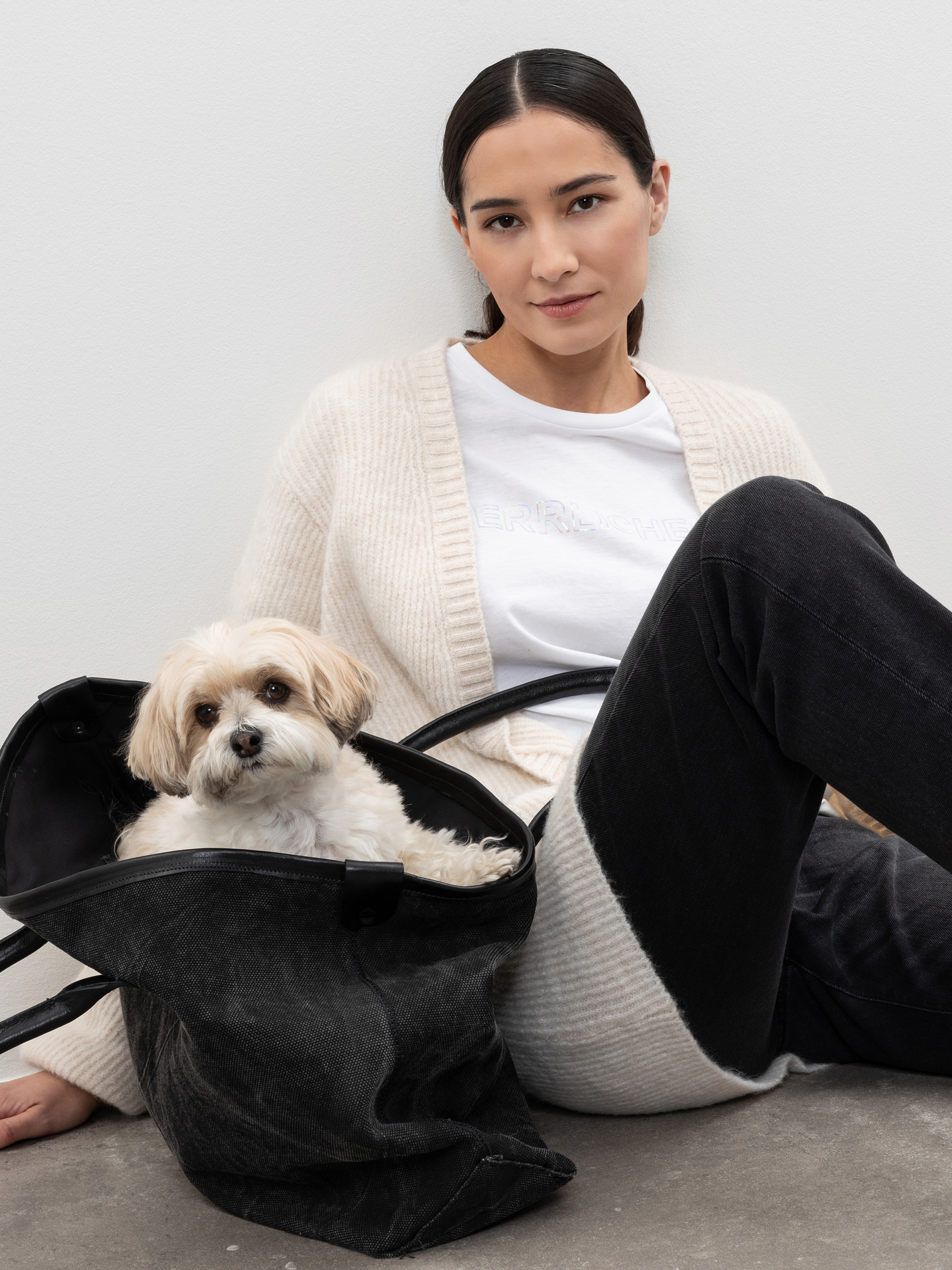 Eine Frau mit dunklem Haar, die einen weißen Pullover und schwarze Jeans trägt, sitzt an einer weißen Wand. Ein kleiner weiß-hellbrauner Hund sitzt in einer schwarzen Tasche neben ihr.