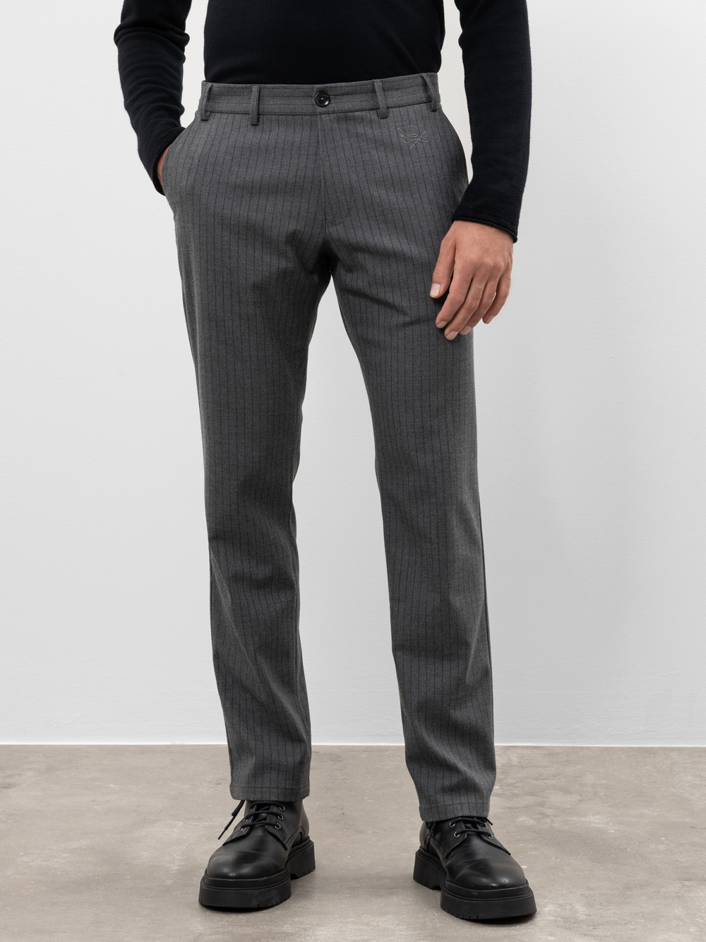 Eine Person, die eine graue Nadelstreifenhose, ein schwarzes langärmeliges Hemd und schwarze Anzugschuhe trägt, steht mit einer Hand in der Tasche vor einer schlichten hellen Wand.