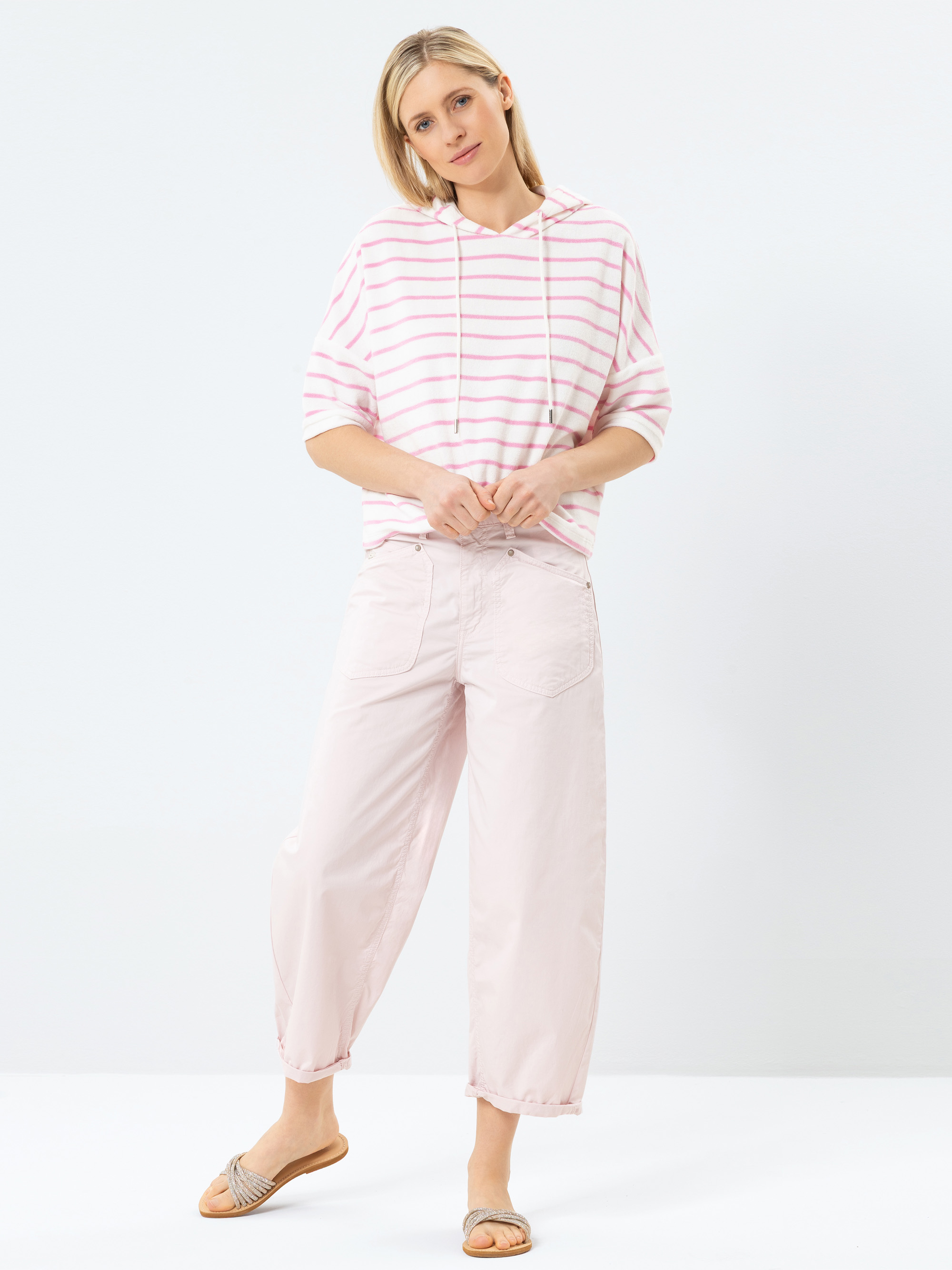 Eine Frau steht vor einem weißen Hintergrund und trägt einen hellrosa und weiß gestreiften Kapuzenpullover, eine hellrosa Hose mit weitem Bein und Slipper-Sandalen. Sie hat blondes Haar und schaut mit einem entspannten Gesichtsausdruck leicht zur Seite