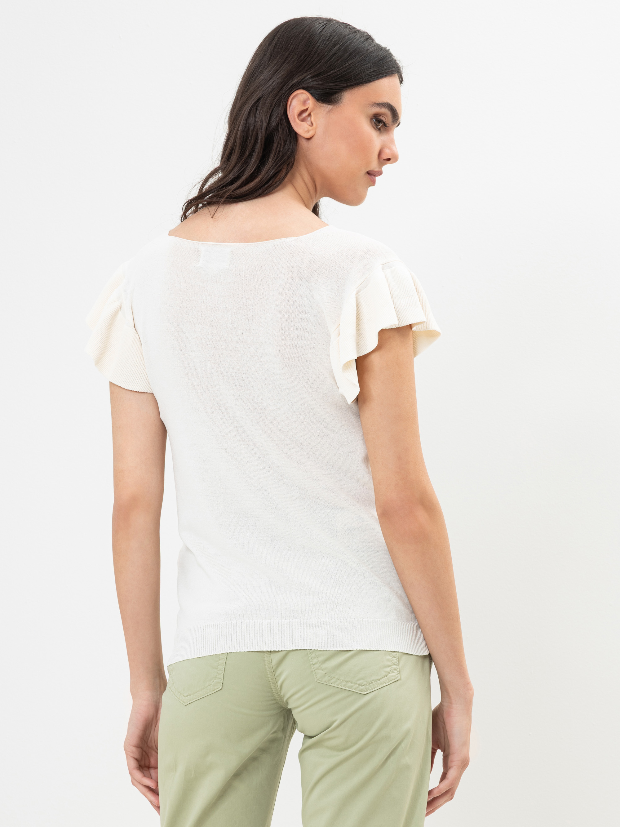 Eine Frau mit langen dunklen Haaren trägt ein weißes kurzärmeliges Oberteil mit gerüschten Ärmeln und eine hellgrüne Hose. Sie steht mit dem Rücken zugewandt und blickt zur Seite vor einem schlichten weißen Hintergrund.