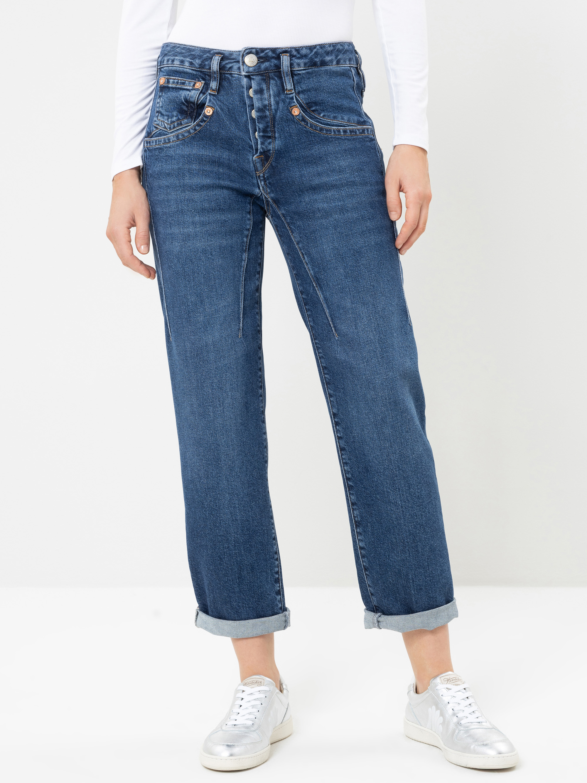 Eine Person, die eine blaue Jeans mit hoher Taille und umgeschlagenen Säumen, ein weißes Langarm-Oberteil und silberne Sneaker trägt und vor einem schlichten, hellen Hintergrund steht.