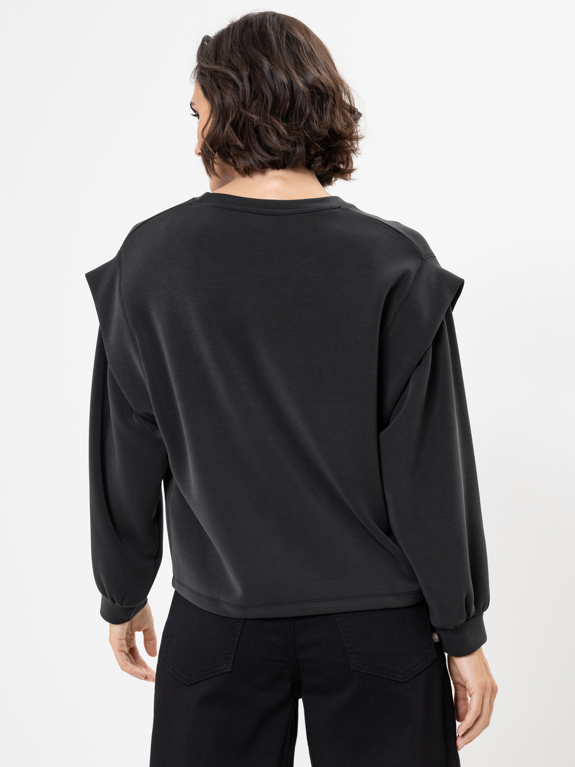 Eine Person mit kurzen, gewellten braunen Haaren wird von hinten gezeigt. Sie trägt ein locker sitzendes schwarzes langärmeliges Oberteil mit leicht überschnittenen Schultern und eine schwarze Hose vor einem schlichten weißen Hintergrund.