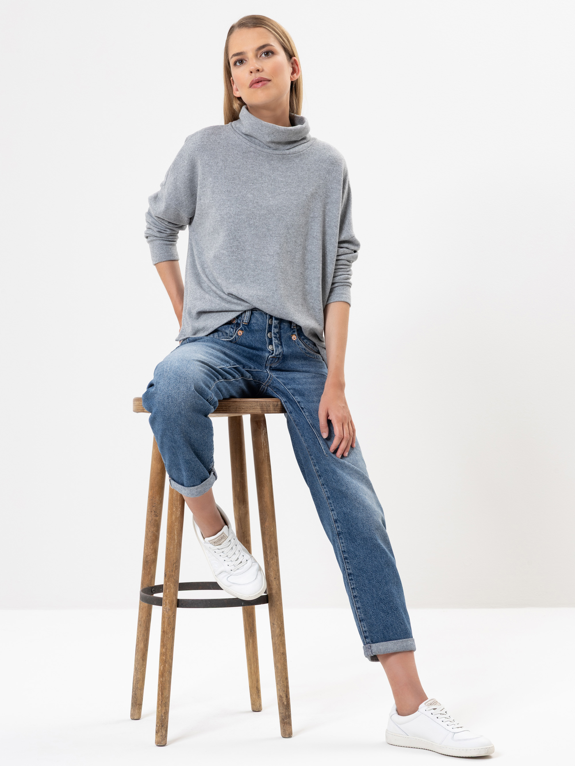 Eine Frau in einem grauen Rollkragenpullover, blauen Jeans und weißen Turnschuhen sitzt auf einem Holzhocker vor einem schlichten weißen Hintergrund und blickt mit entspannter Miene leicht nach oben.