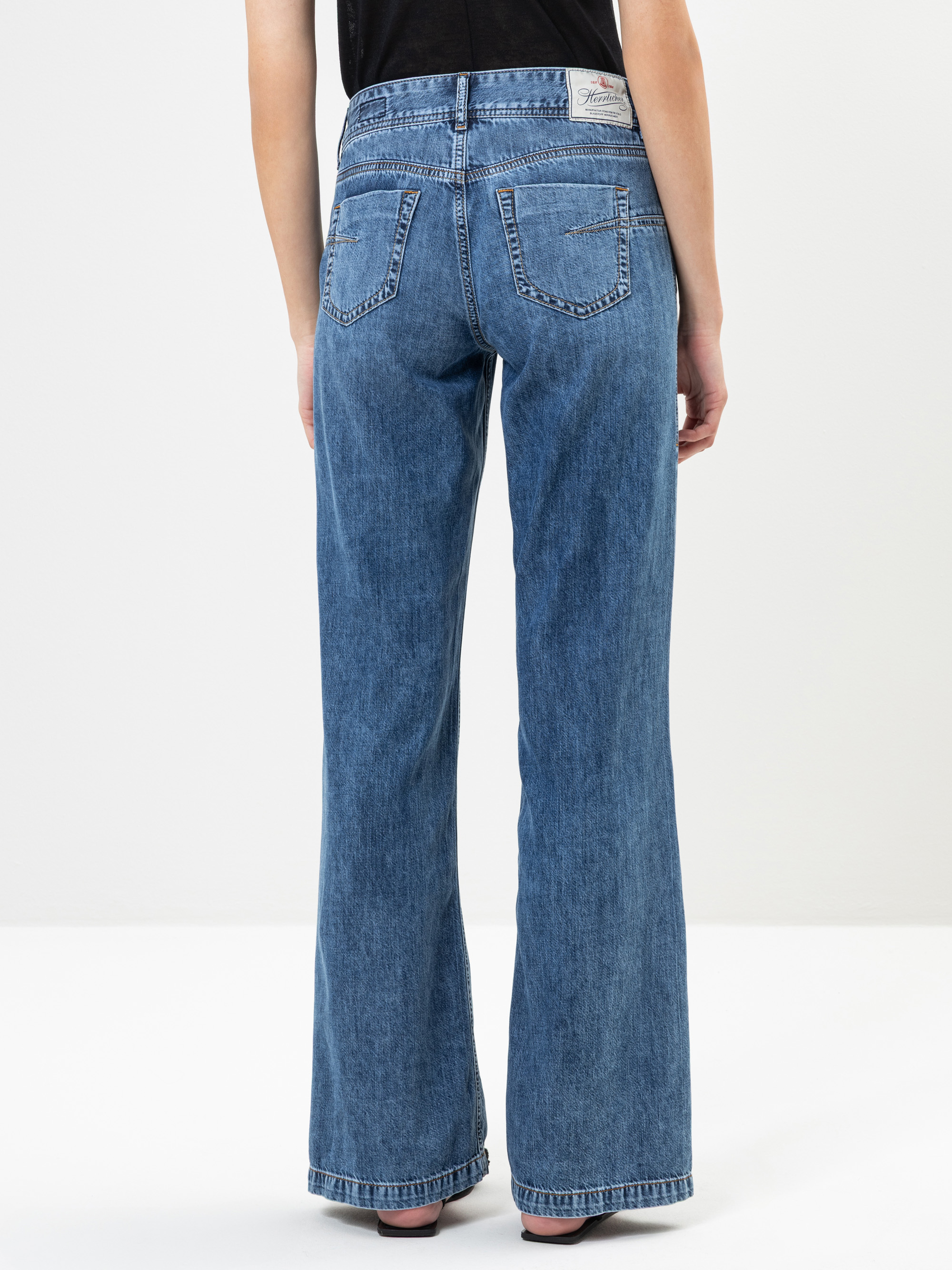 Eine Person, die eine blaue Jeans mit weitem Bein und ein schwarzes Oberteil trägt, ist von hinten zu sehen und steht barfuß vor einem schlichten weißen Hintergrund.