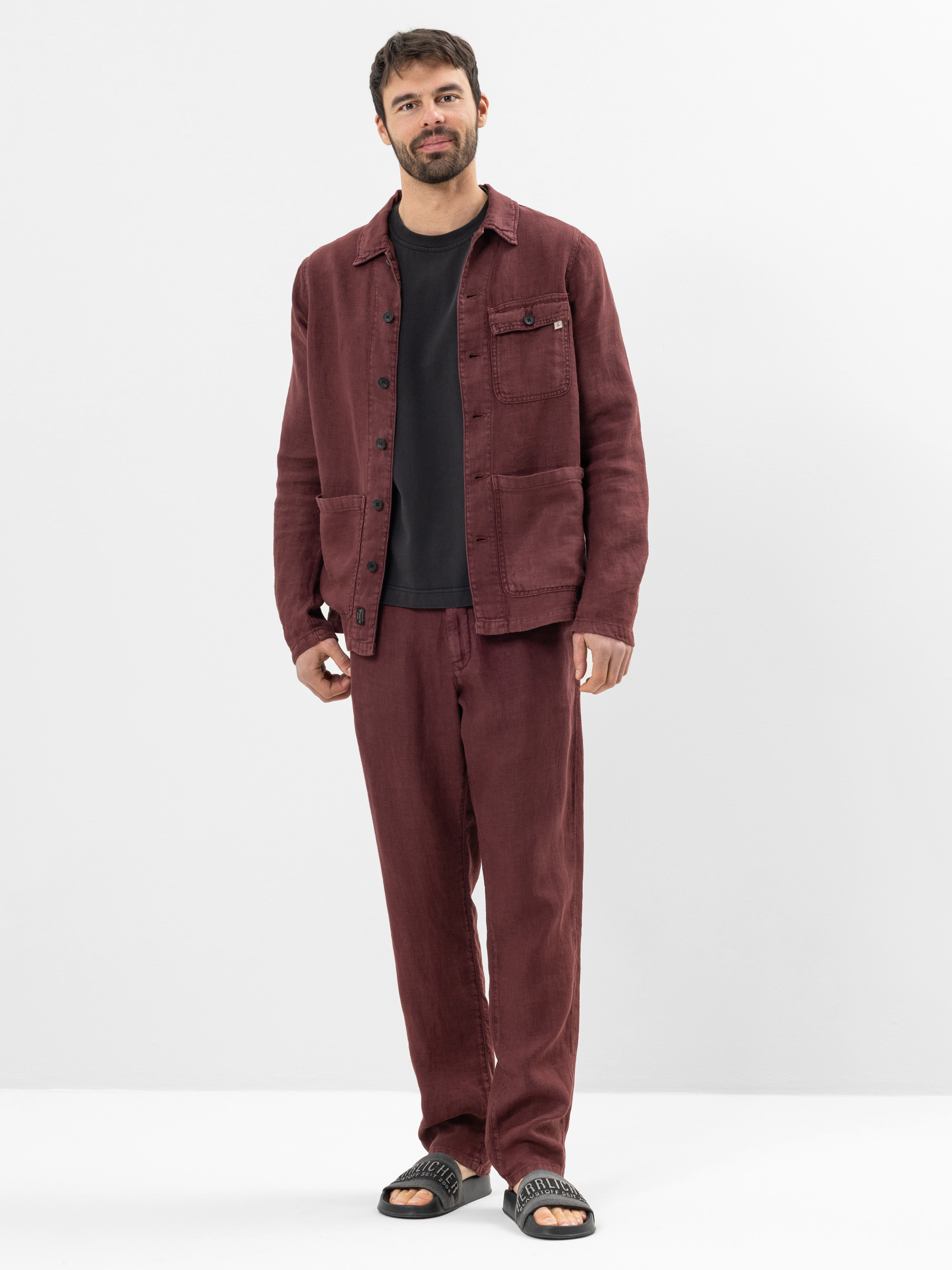 Ein Mann steht vor einem schlichten weißen Hintergrund und trägt eine passende kastanienbraune Jacke und Hose, ein schwarzes T-Shirt und schwarze Slide-Sandalen. Er hat kurzes dunkles Haar, einen Bart und einen entspannten, lässigen Gesichtsausdruck.