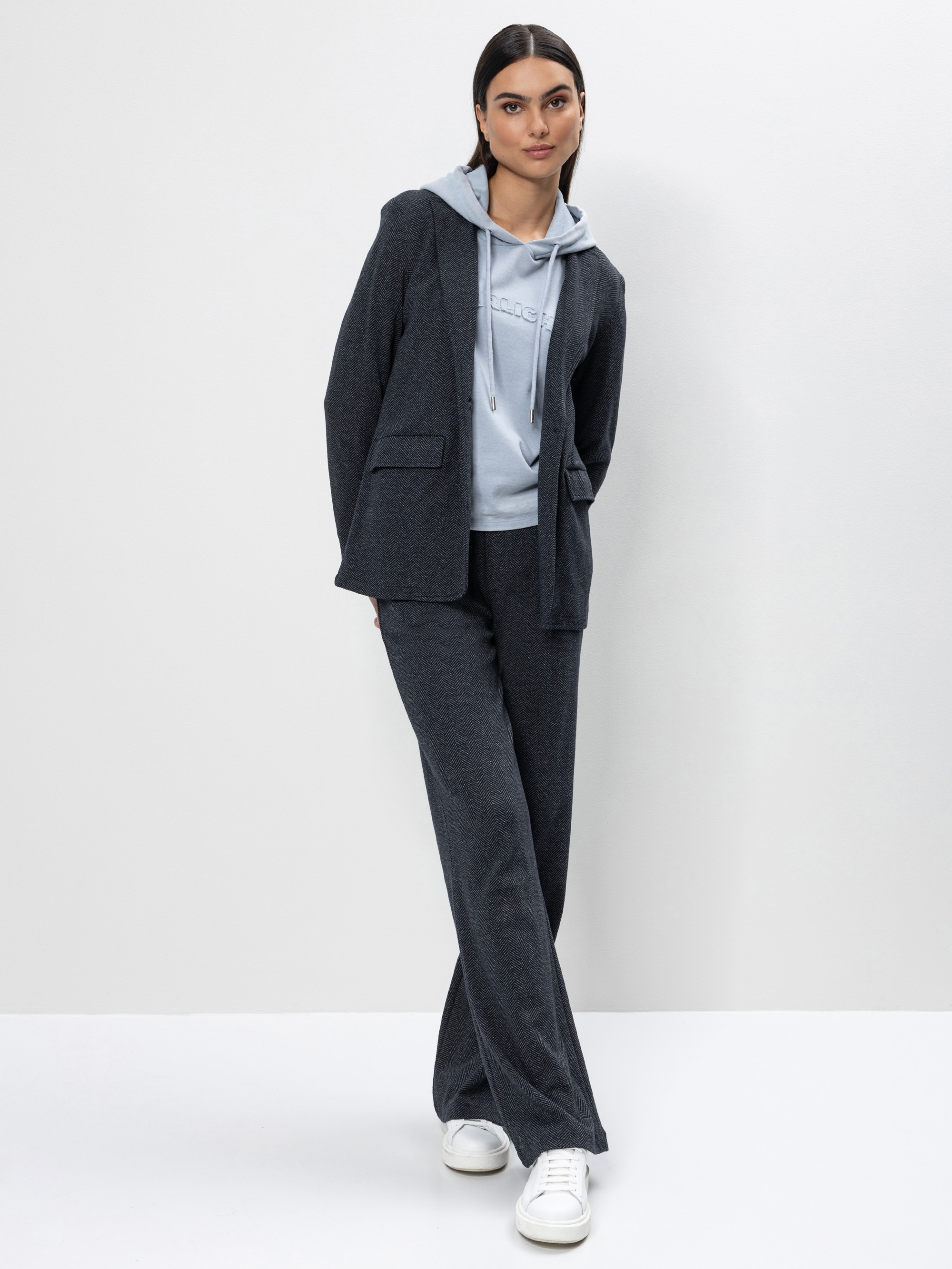 Eine Frau steht vor einem weißen Hintergrund und trägt einen dunkelgrauen Blazer und eine Hose über einem hellblauen Kapuzenpullover, dazu weiße Turnschuhe. Sie hat glattes dunkles Haar und einen neutralen Gesichtsausdruck, eine Hand steckt in ihrer T