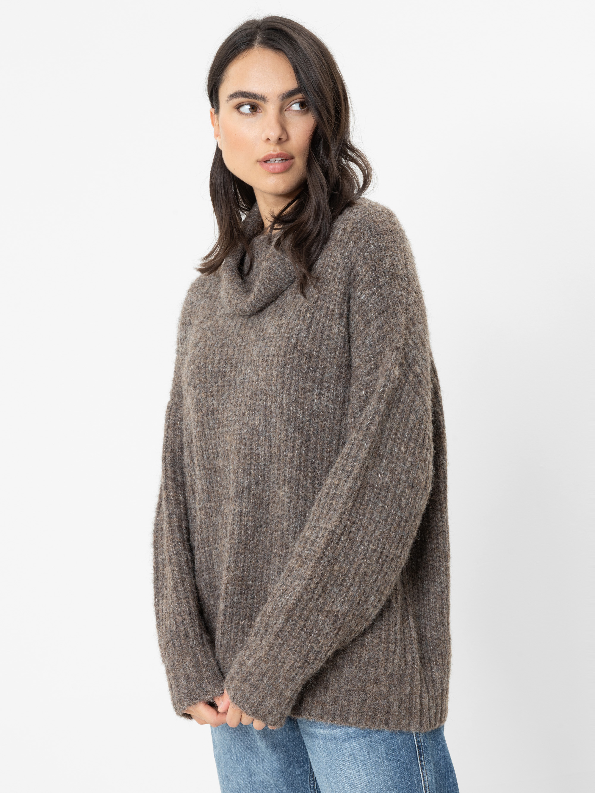 Eine Frau mit dunklem Haar trägt einen lockeren, strukturierten braunen Pullover und blaue Jeans. Sie steht vor einem schlichten weißen Hintergrund und blickt mit ausdruckslosem Gesicht nach links.