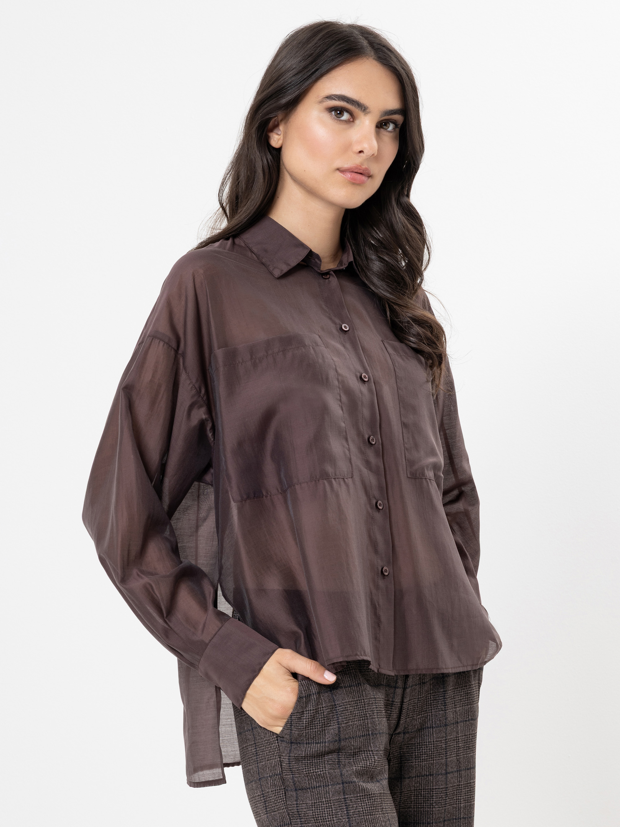 Eine Frau mit langen dunklen Haaren, die ein lockeres, durchsichtiges braunes Button-up-Hemd und eine karierte Hose trägt, steht mit einer Hand in der Tasche vor einem schlichten weißen Hintergrund.