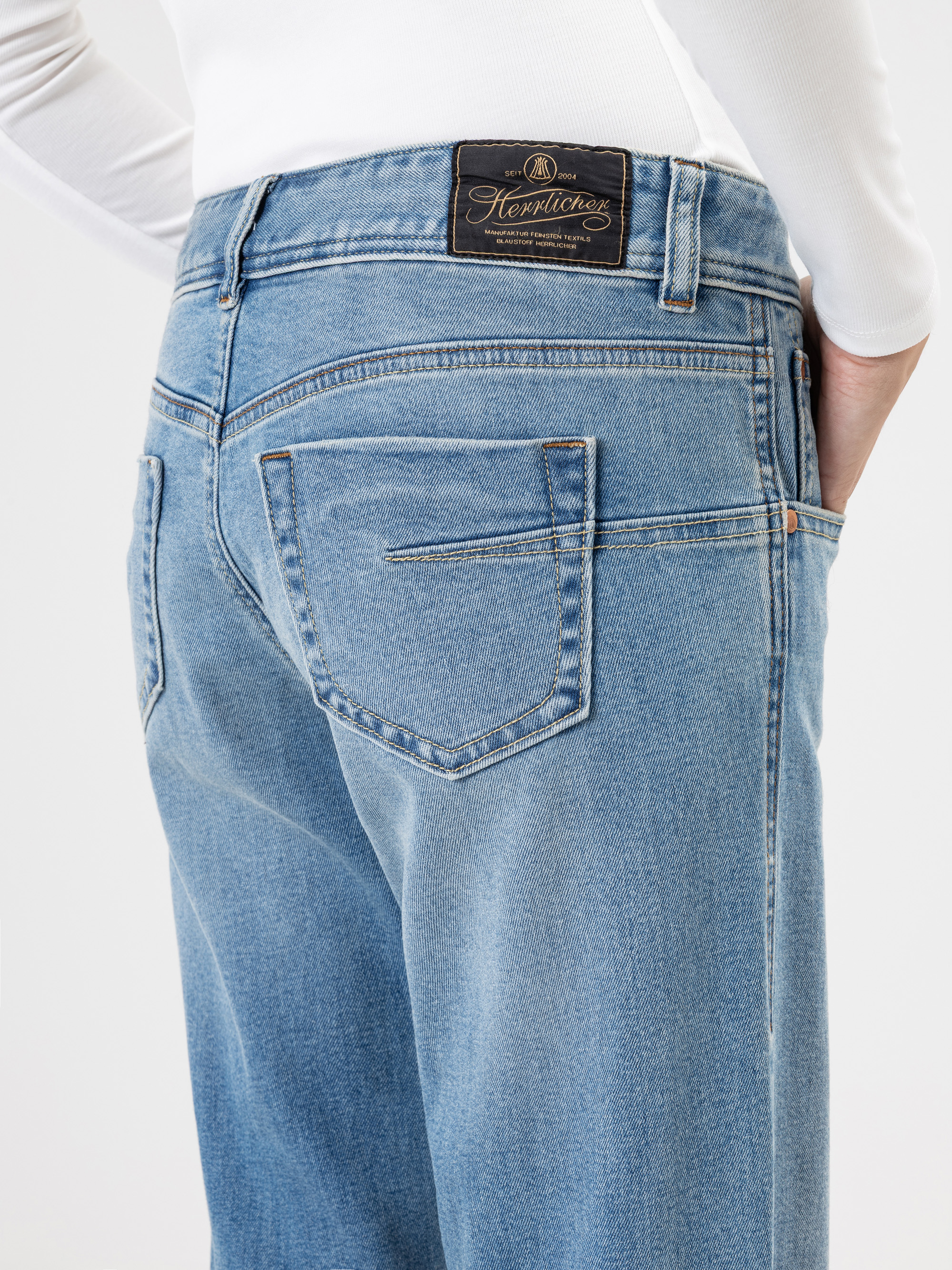 Person, die eine hellblaue Jeans mit hoher Taille und ein weißes langärmeliges Hemd trägt und eine Hand in der Gesäßtasche hat. Die Jeans hat einen braunen Lederaufnäher mit einem Markenlogo über der rechten Tasche.