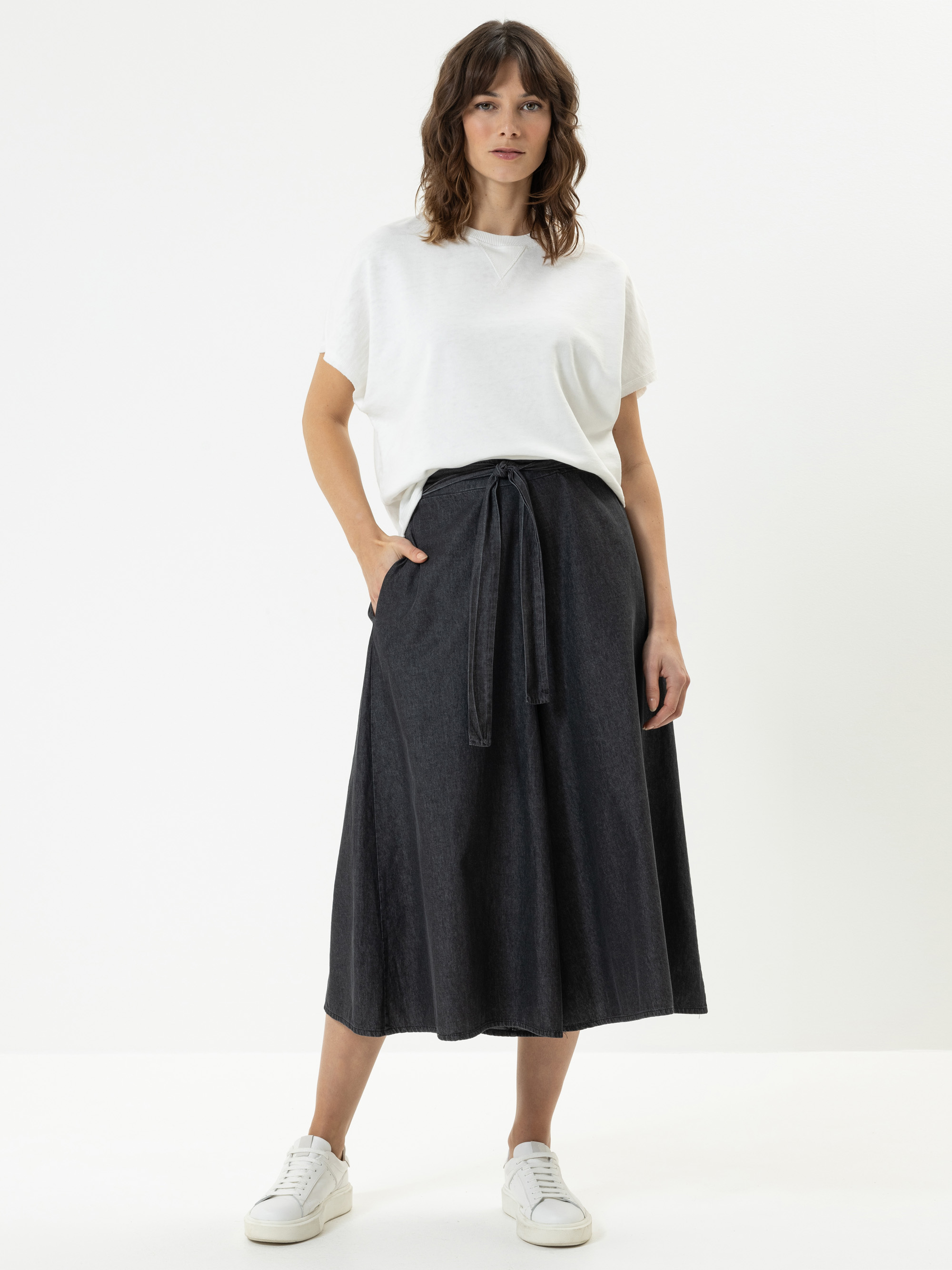 Eine Frau steht vor einem weißen Hintergrund und trägt ein weißes, kurzärmeliges T-Shirt, einen schwarzen, hochtaillierten Midirock mit Bindegürtel und weiße Turnschuhe. Sie hat eine Hand in der Tasche und einen neutralen Gesichtsausdruck.