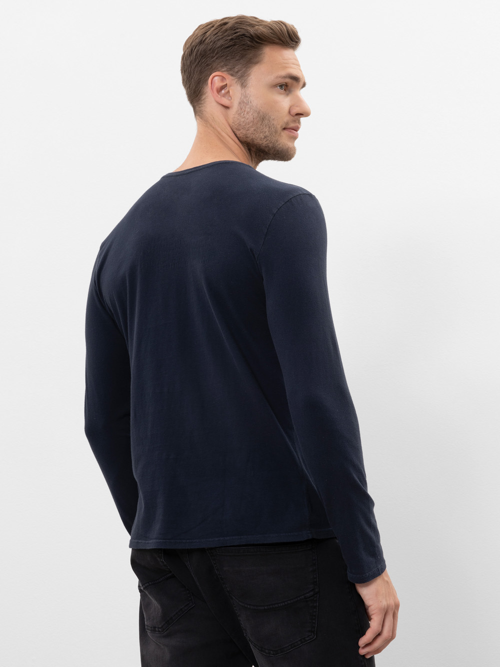 Ein Mann mit kurzen braunen Haaren steht mit dem Gesicht zur Seite und schaut über seine Schulter. Er trägt ein langärmeliges, dunkles, marineblaues Hemd und schwarze Jeans vor einem schlichten weißen Hintergrund.