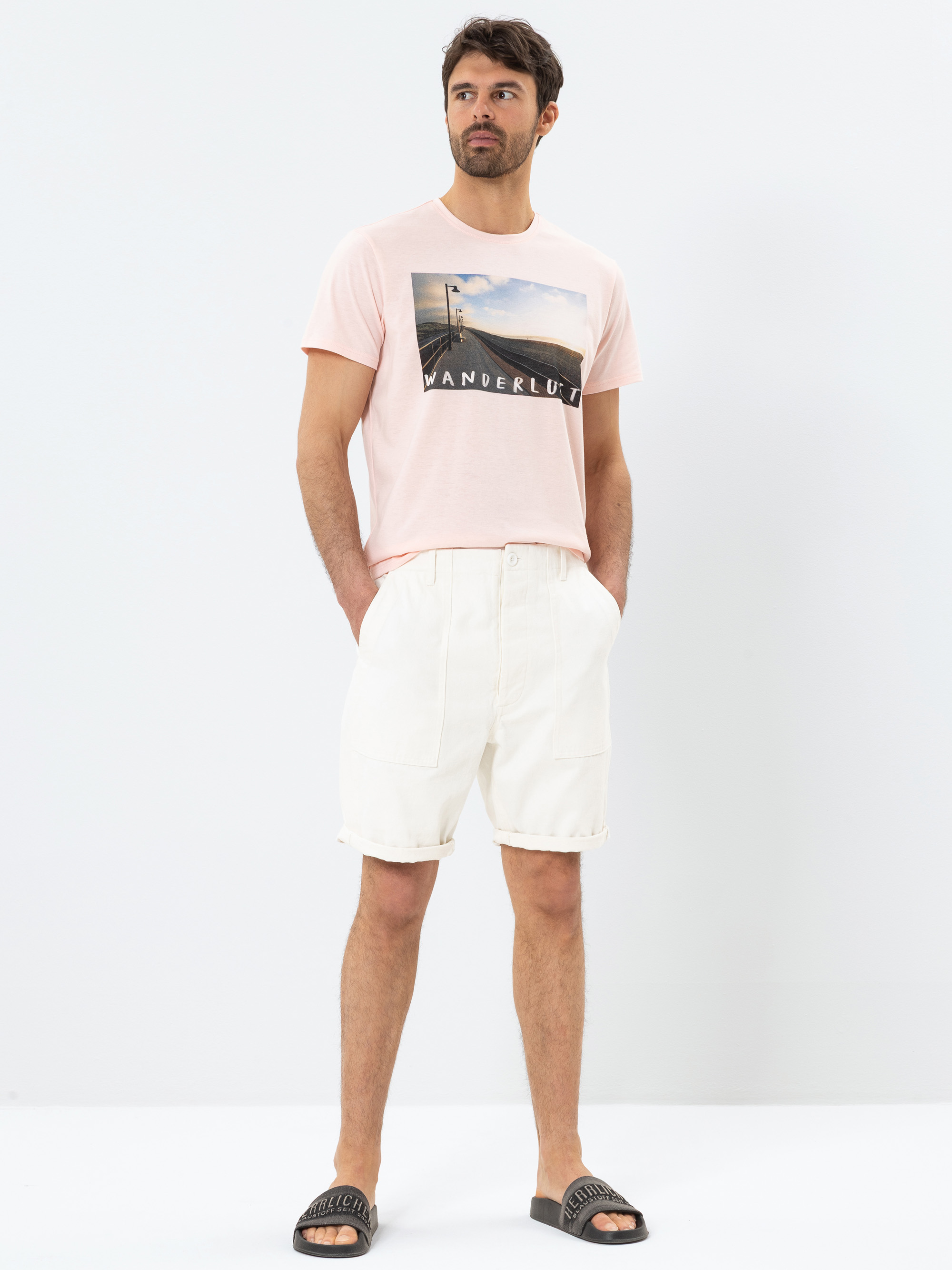Ein Mann, der ein hellrosa T-Shirt mit Wanderlust-Grafik, cremefarbene Shorts und schwarze Slide-Sandalen trägt, steht vor einem schlichten weißen Hintergrund, hat die Hände in den Taschen und schaut leicht zur Seite.