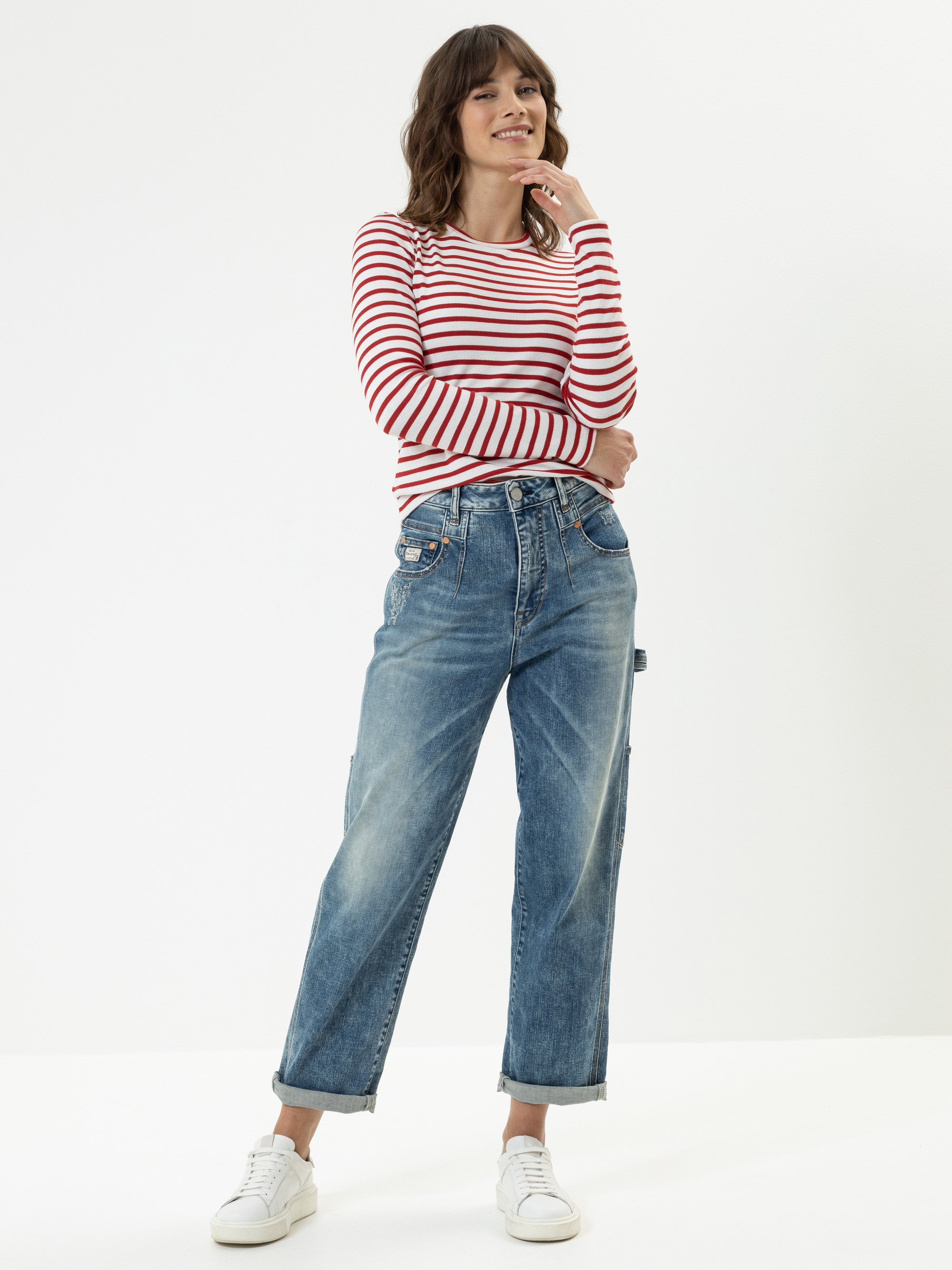 Eine Frau steht lächelnd vor einem schlichten weißen Hintergrund. Sie trägt ein rot-weiß gestreiftes, langärmeliges Hemd, weite blaue Jeans mit aufgerollten Manschetten und weiße Turnschuhe. Ihre Arme sind verschränkt und sie hat gewelltes braunes