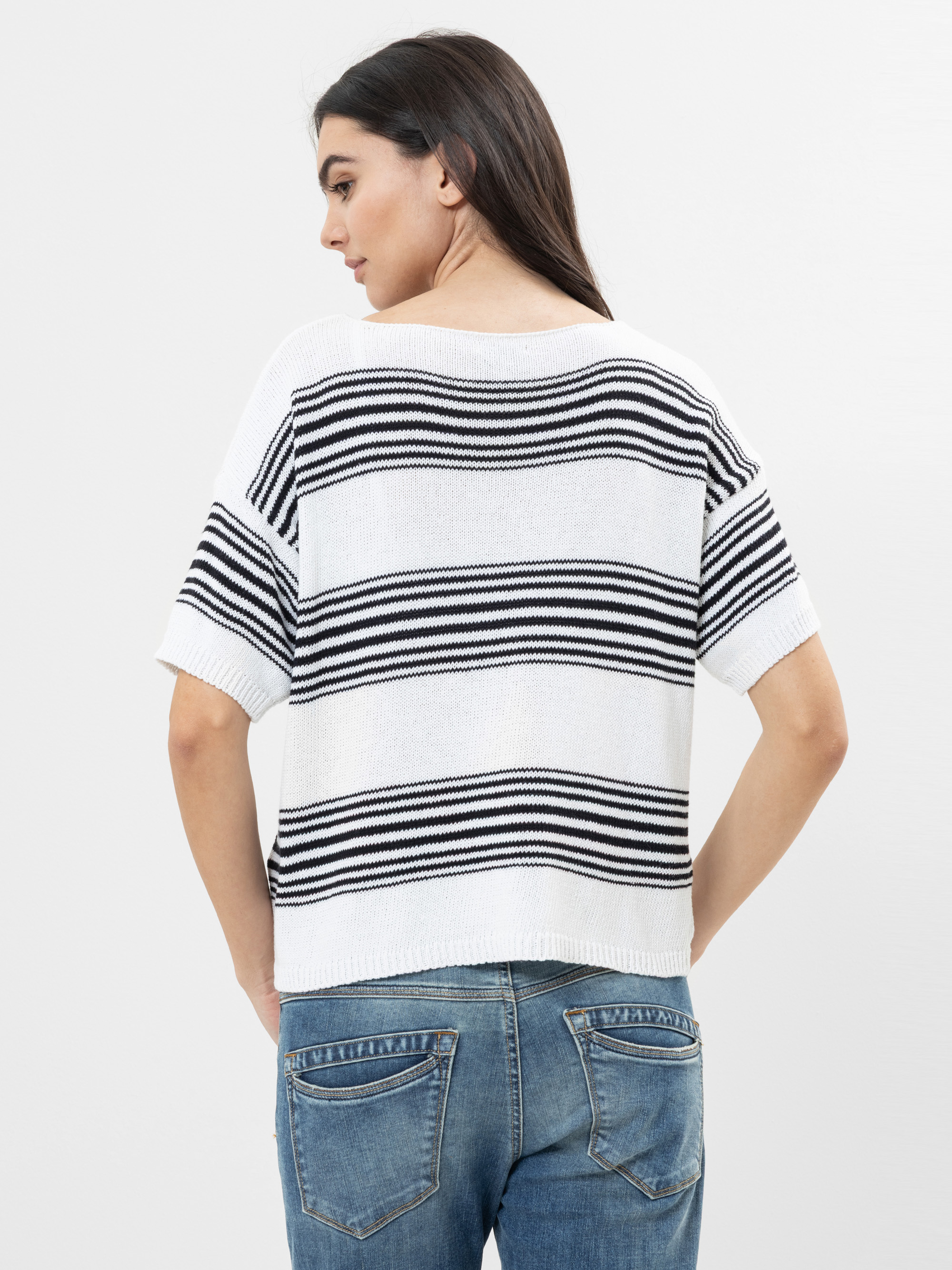 Eine Frau mit langen dunklen Haaren, die einen weißen kurzärmeligen Pullover mit schwarzen Querstreifen und blaue Jeans trägt, ist von hinten vor einem schlichten hellen Hintergrund zu sehen.