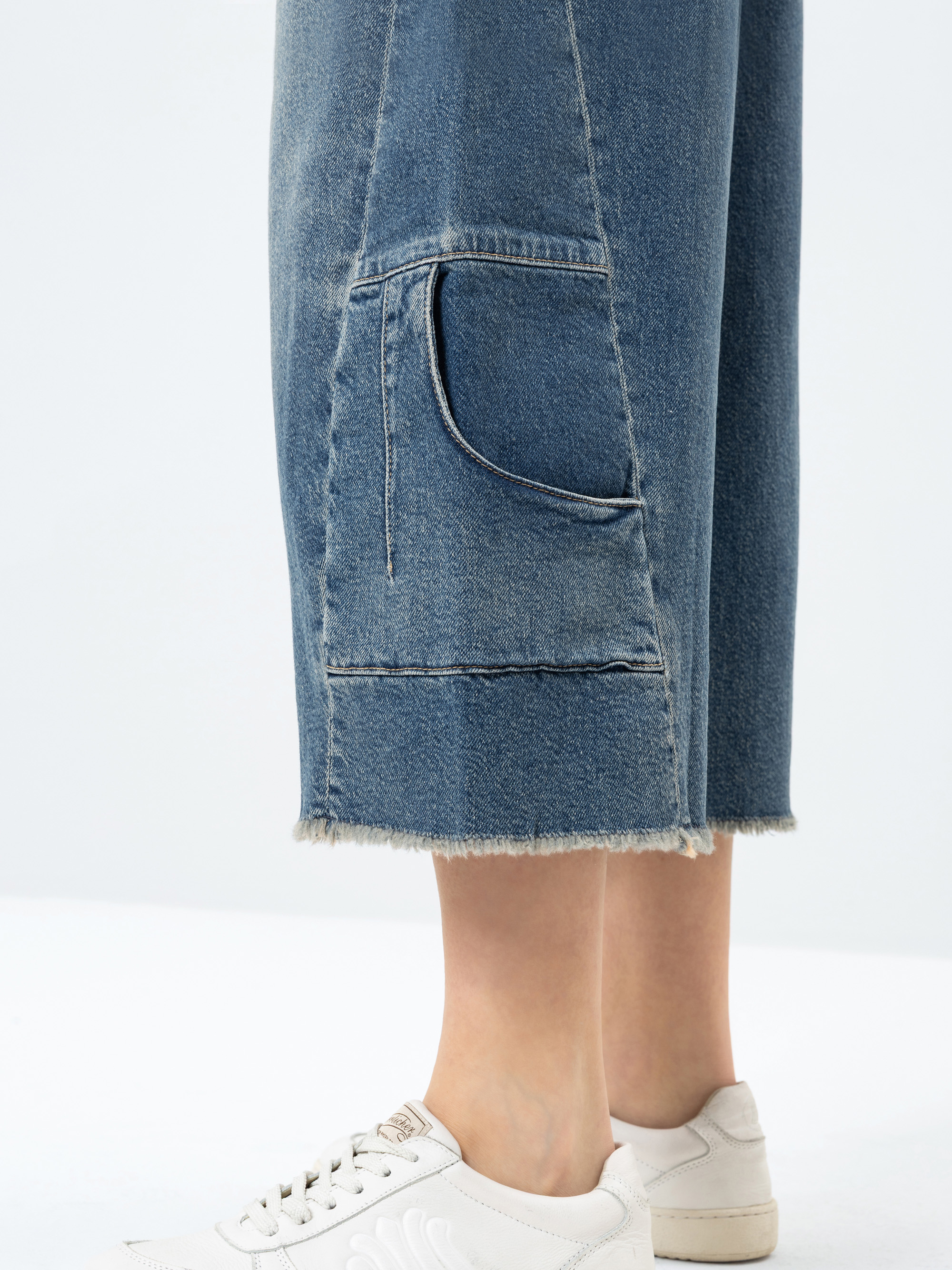 Eine Person trägt eine hellblaue abgeschnittene Jeanshose mit ausgefransten Säumen und einer geschwungenen Vordertasche, gepaart mit weißen Turnschuhen. Nur die Unterschenkel und Füße sind vor einem einfarbigen Hintergrund zu sehen.
