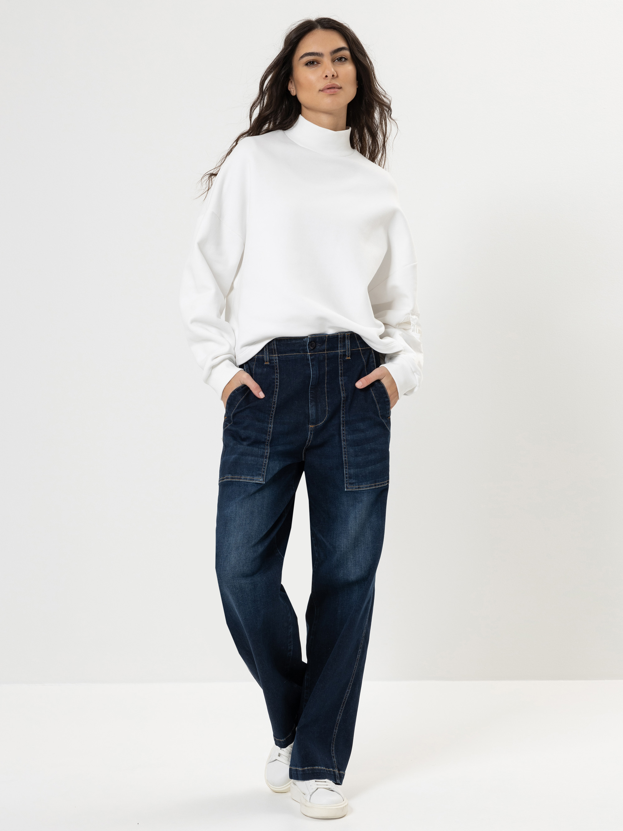 Eine Person steht vor einem einfarbig weißen Hintergrund und trägt ein weißes Rollkragen-Sweatshirt, dunkle Jeans mit weitem Bein und großen Vordertaschen sowie weiße Turnschuhe. Ihre Hände stecken in den Taschen und sie hat langes, gewelltes Haar.
