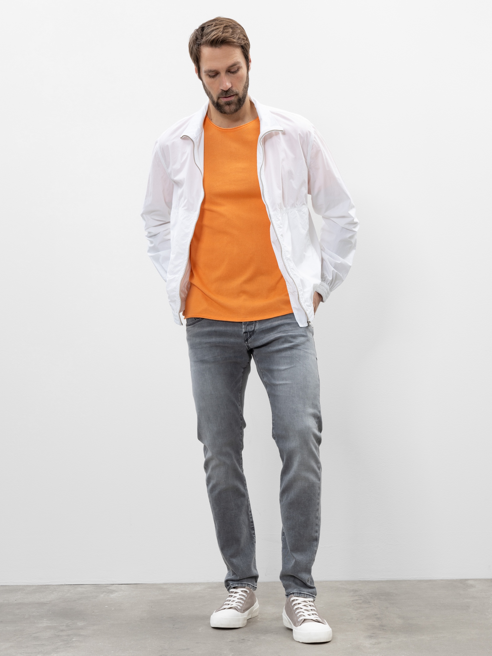Ein Mann steht vor einem schlichten weißen Hintergrund und trägt ein leuchtend orangefarbenes Hemd, eine weiße Jacke, graue Jeans und weiße Turnschuhe. Seine Hände stecken in den Jackentaschen und er blickt nach unten.
