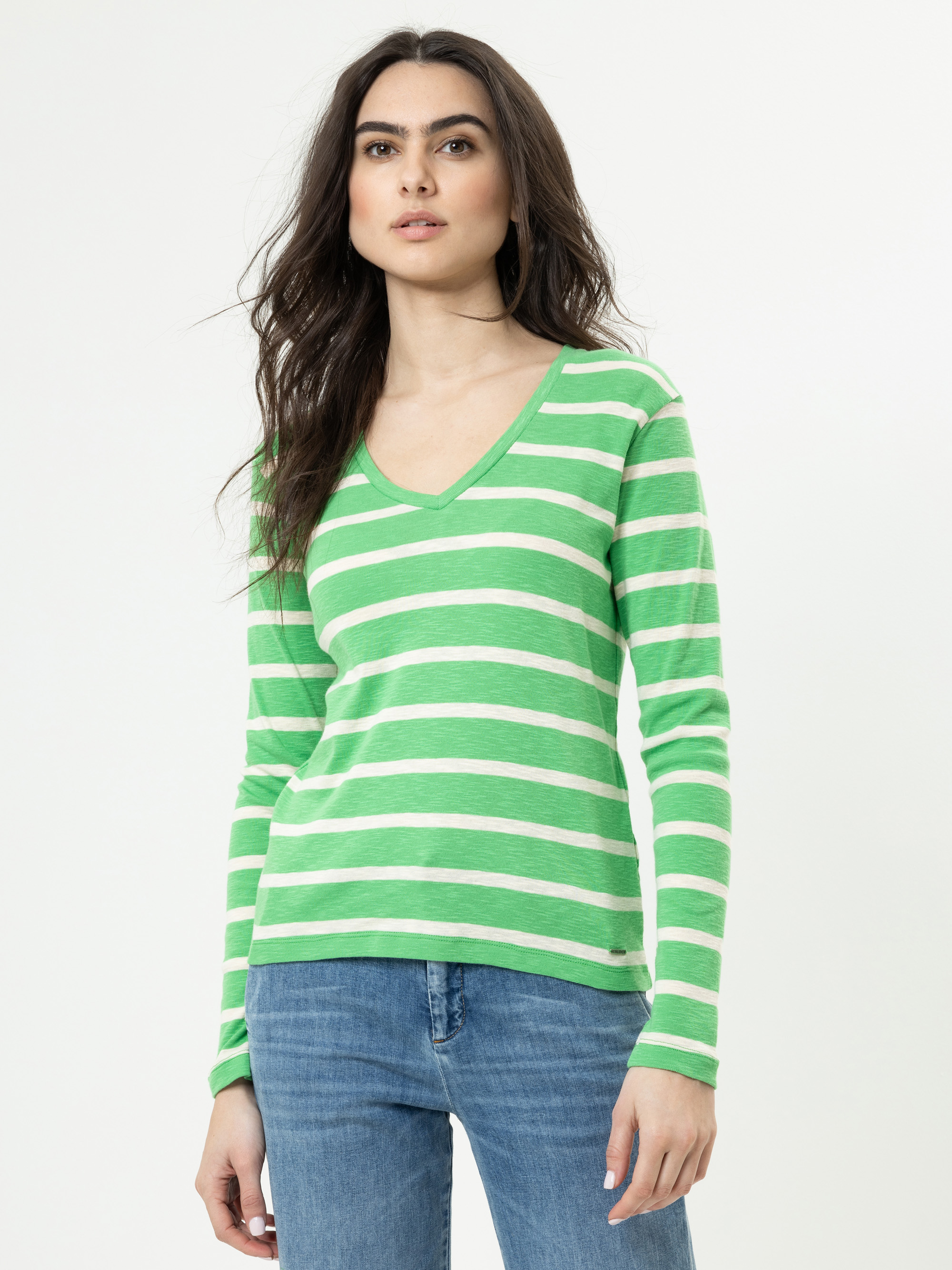 Eine Person mit langen braunen Haaren trägt ein grün-weiß gestreiftes, langärmeliges Hemd mit V-Ausschnitt und blaue Jeans und steht vor einem schlichten weißen Hintergrund.