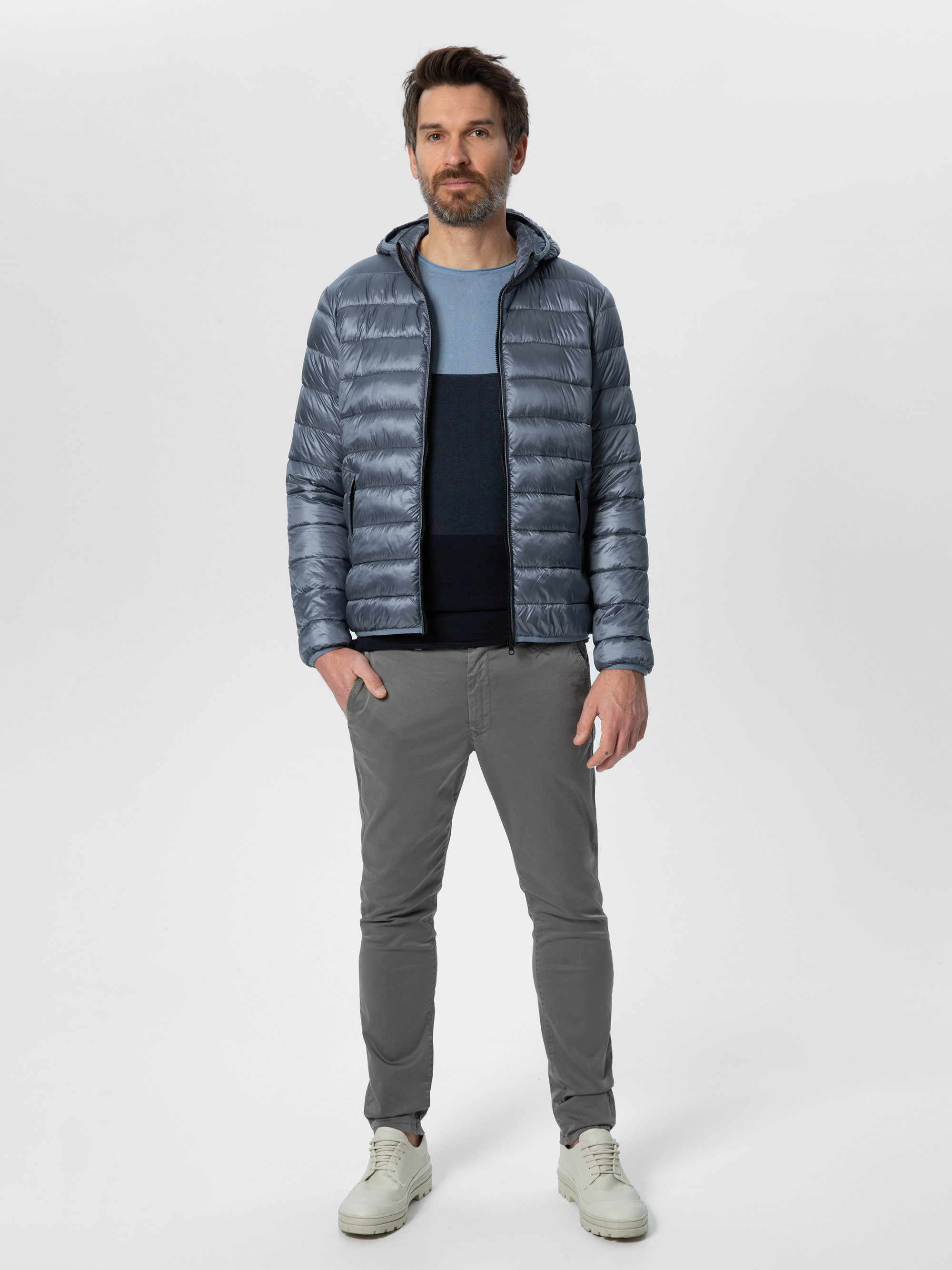 Ein Mann mit kurzen dunklen Haaren und einem Bart steht mit dem Gesicht nach vorne und trägt eine glänzende graue Pufferjacke, einen blauen Pullover, eine graue Hose und weiße Turnschuhe vor einem schlichten weißen Hintergrund.