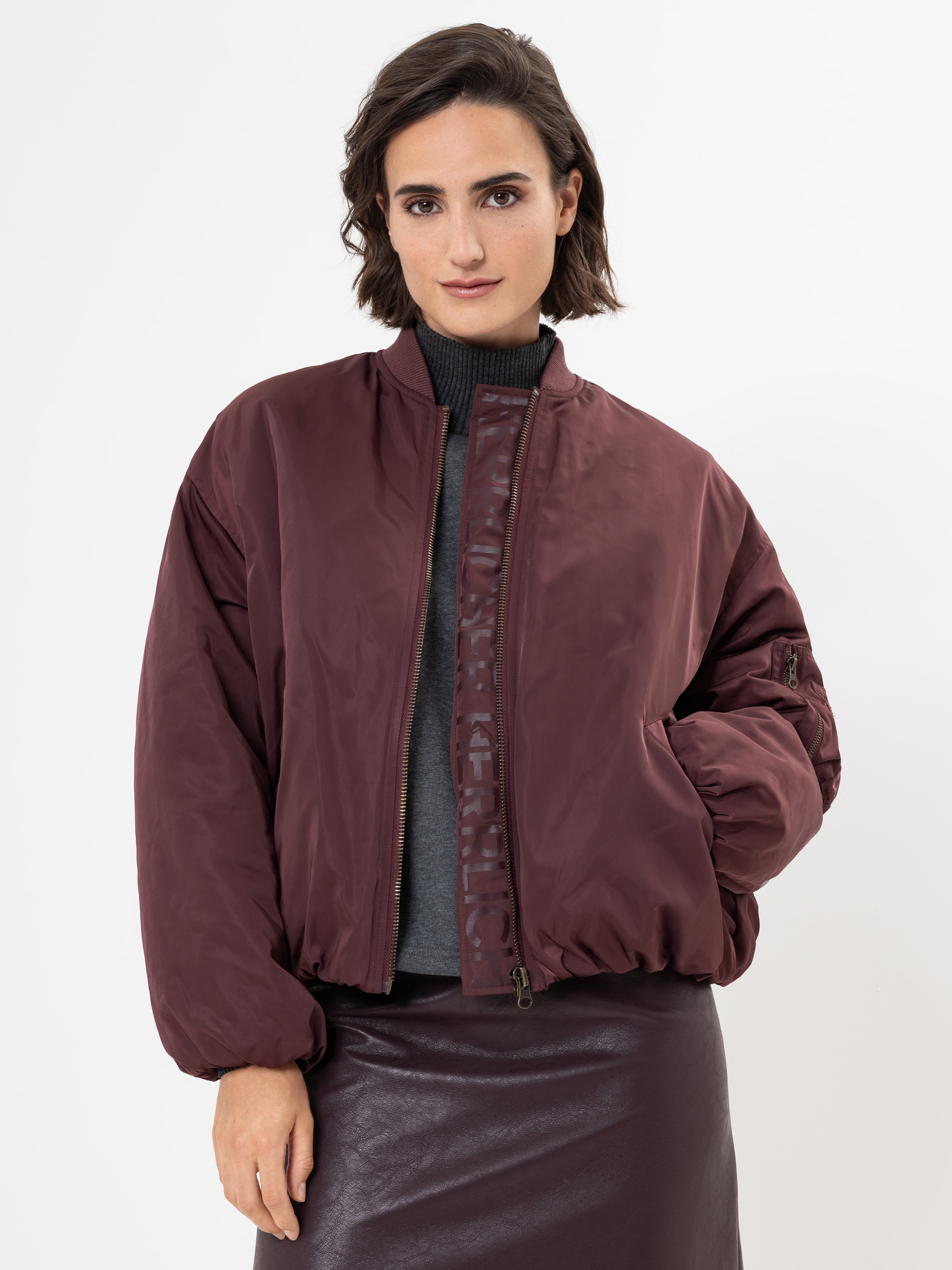 Eine Frau mit kurzen braunen Haaren trägt eine kastanienbraune Bomberjacke über einem grauen Rollkragenpullover und einen dunkelbraunen Lederrock. Sie steht vor einem schlichten weißen Hintergrund und hat eine Hand in der Tasche.