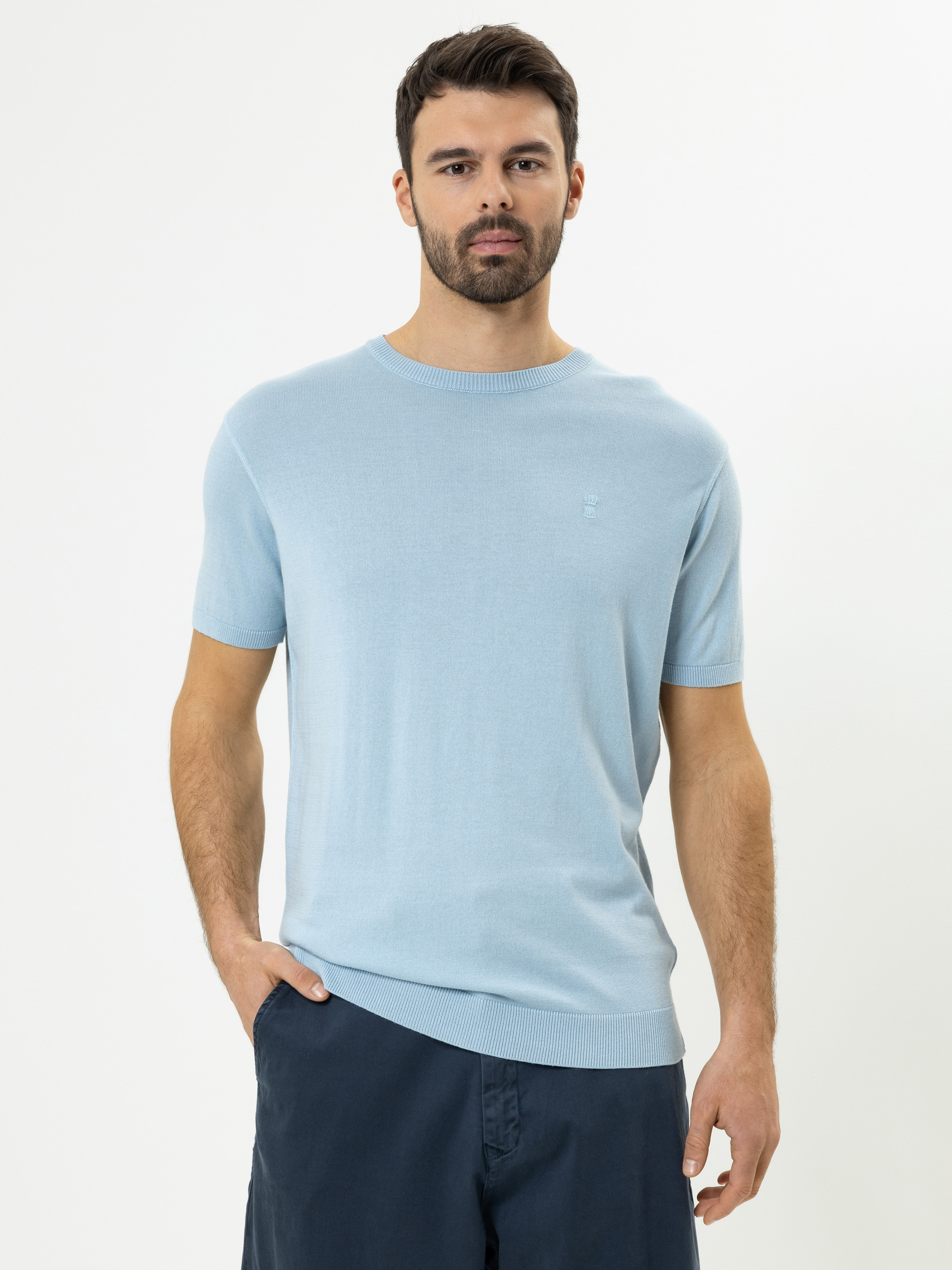 Ein Mann mit dunklem Haar und Bart trägt einen hellblauen kurzärmeligen Pullover und dunkelblaue Shorts und steht vor einem schlichten weißen Hintergrund.